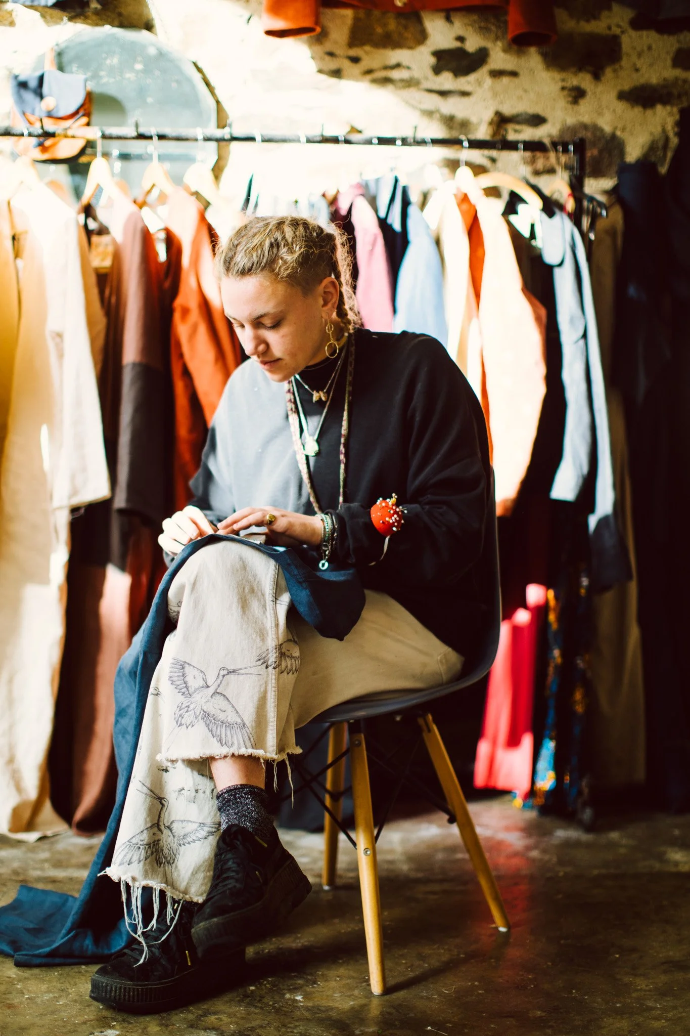A woman with braided hair sitting on a black chair in a vintage clothing store, surrounded by colorful hanging clothes and a stone wall. She is wearing a black sweatshirt, beige pants with bird embroidery, and black platform shoes, focusing on sewing
