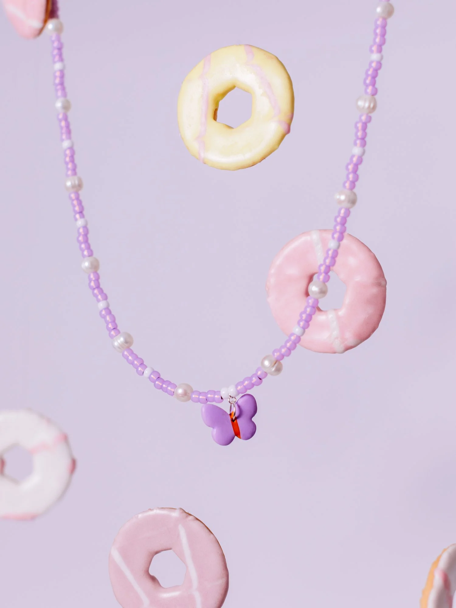 Purple beaded necklace with a purple butterfly pendant, decorated with pastel-colored donuts in pink, yellow, and white, hanging against a light purple background.