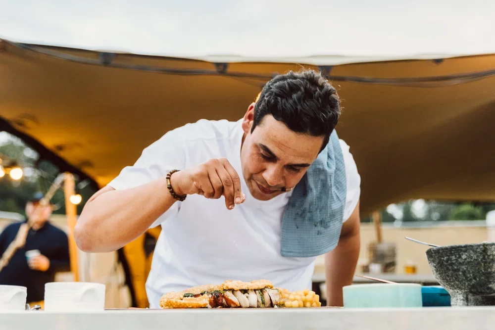 Male chef garnishing a dish at an outdoor event with a tent and blurred background.