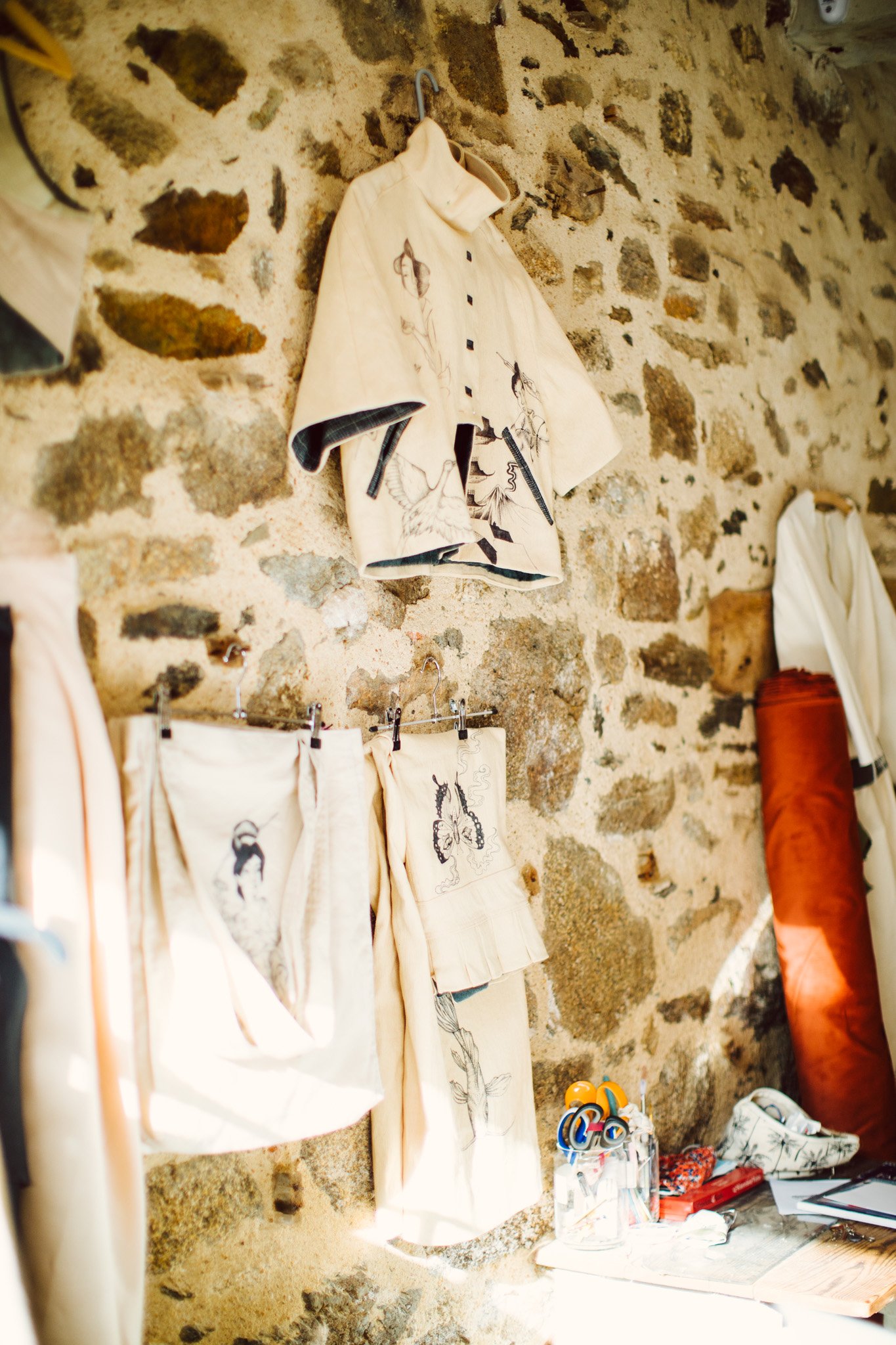 A stone wall with hanging clothing and fabrics featuring embroidered illustrations of people and animals. A desk with stationery and objects is below.