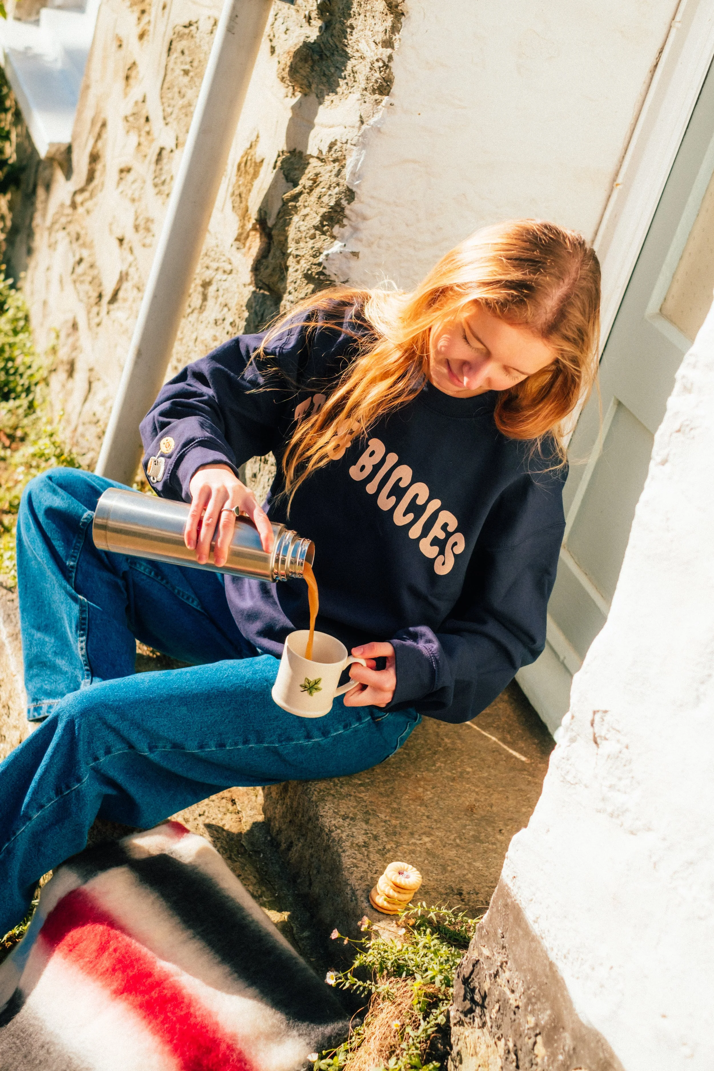 A young woman with red hair, wearing a navy blue sweatshirt with 'BICYCLES' printed on it, is sitting outside on a stone step, pouring coffee into a mug with a green leaf design. She is wearing blue jeans and sitting on a red, black, and white blanke