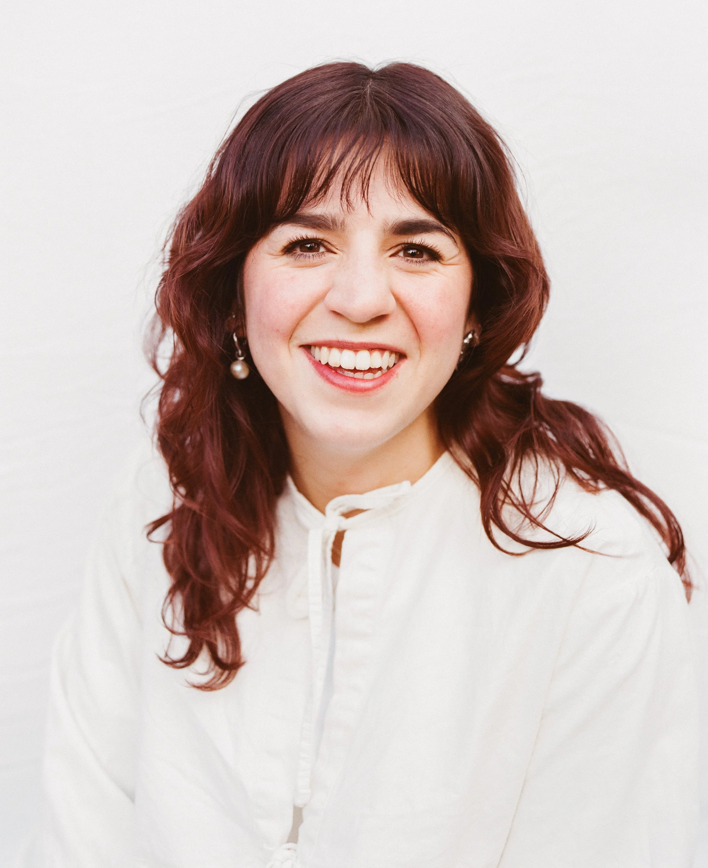 A woman with shoulder-length reddish-brown hair smiling, wearing pearl earrings and a white blouse with a tie at the collar, against a plain white background.
