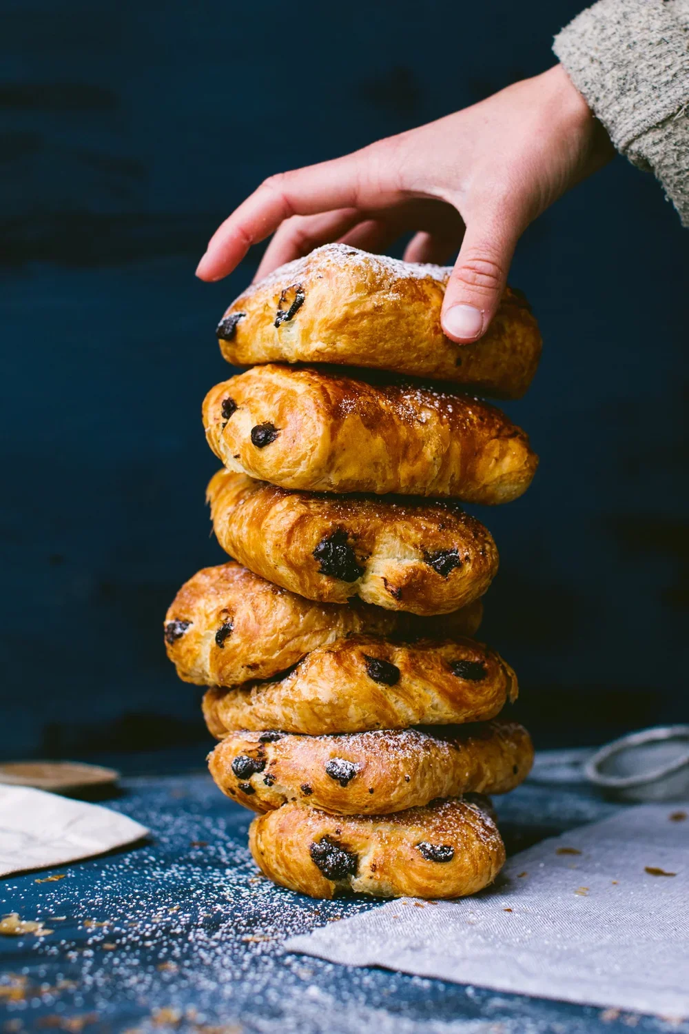 A hand stacking six chocolate chip scones with powdered sugar on top against a dark background.