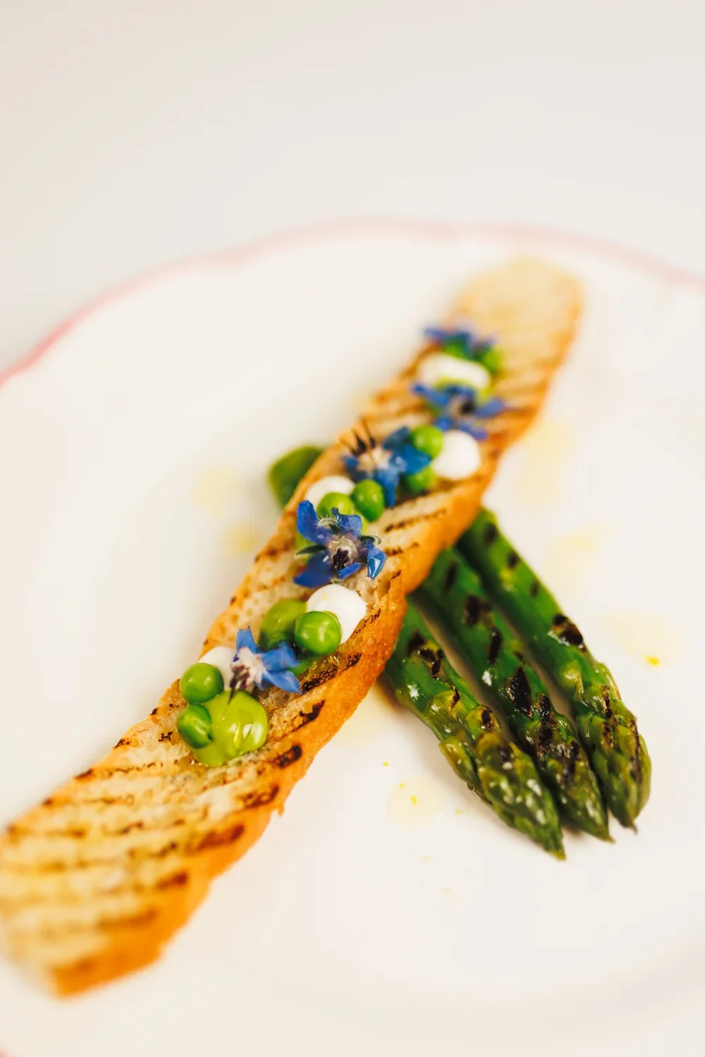 Grilled asparagus spears and a grilled bread slice topped with green peas, edible flowers, and small dollops of white and green sauces.