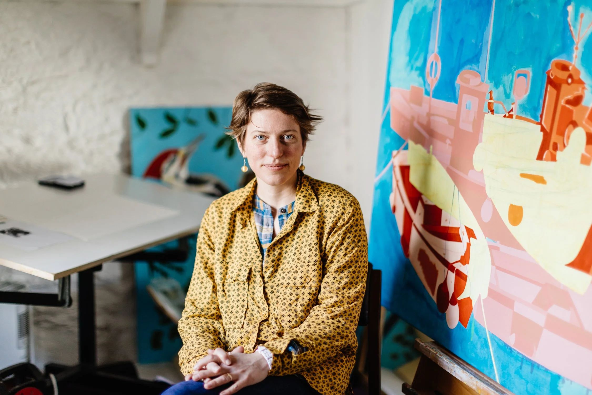A woman with short brown hair, wearing a yellow patterned jacket and earrings, sitting in an art studio with colorful abstract paintings, including a large blue painting and another with a boat, in the background.