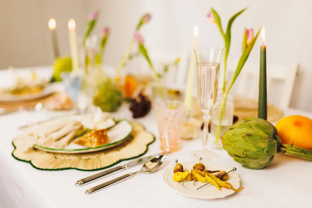 Table setting with a plate of sliced bread and cheese, tall candles with pink flowers, glasses of pink beverage, artichoke, and various fruits like mango and orange.