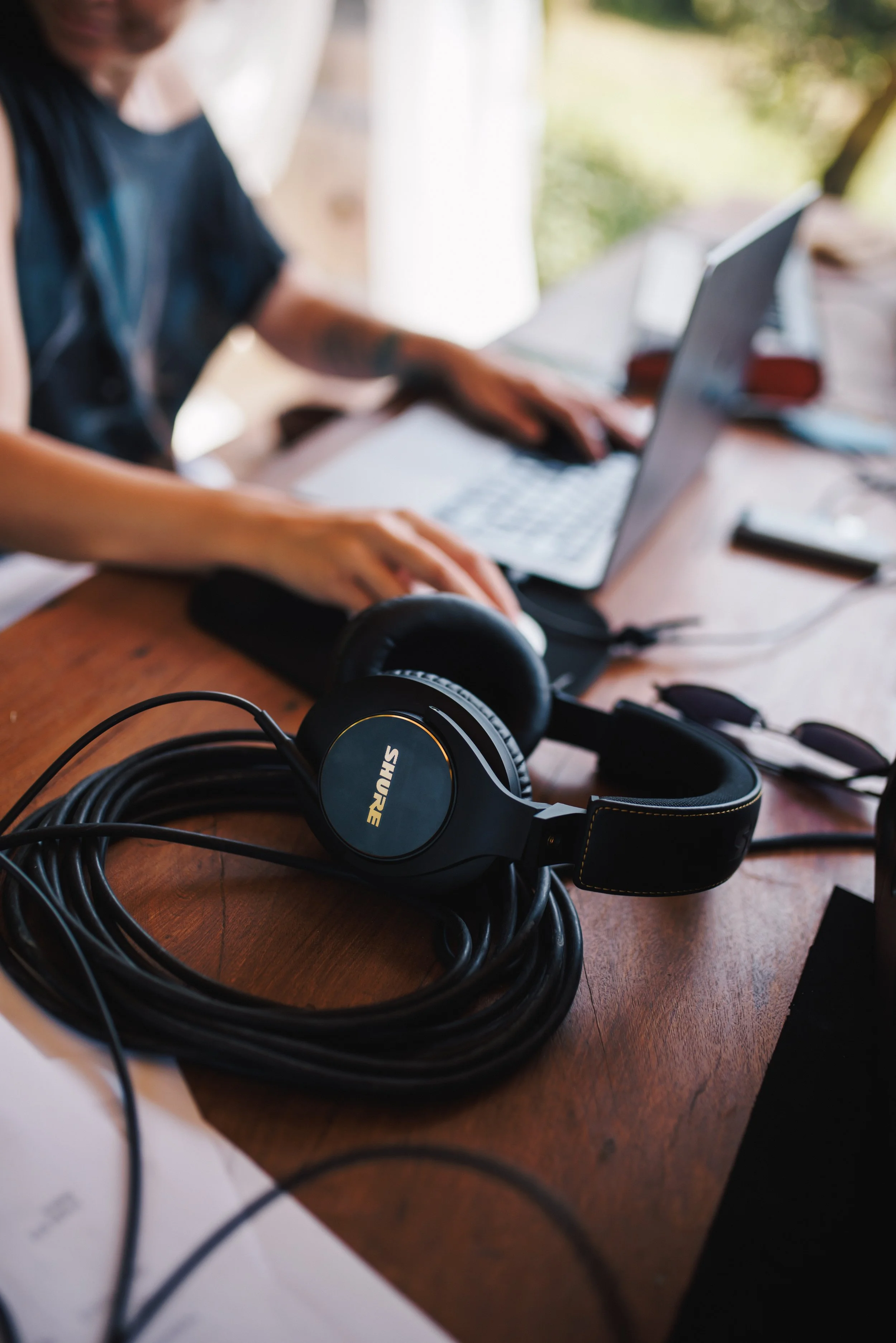 Close-up of black SHURE headphones with a coiled cable on a wooden table, with a person working on a laptop in the background.