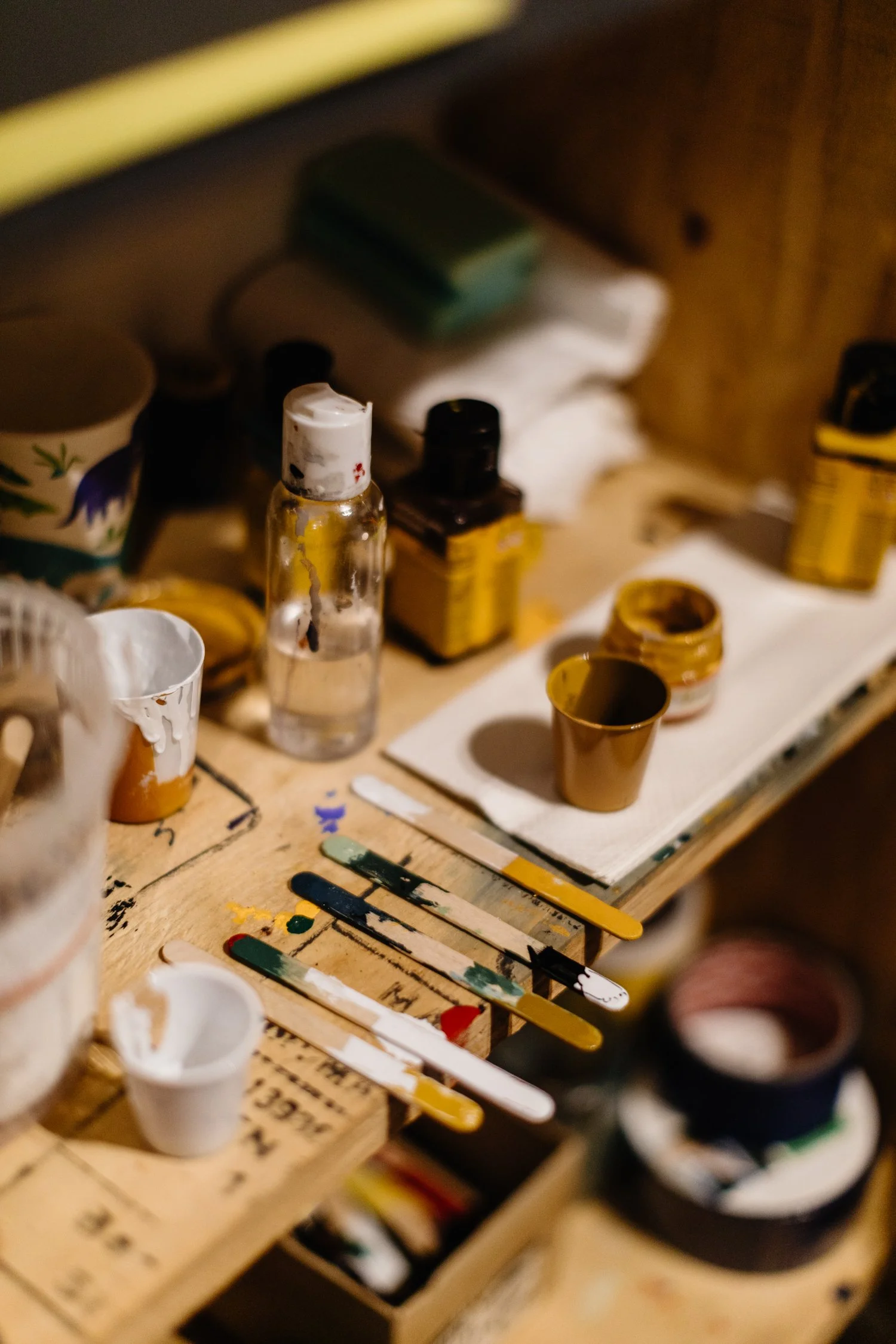 An artist's workspace showing paint bottles, brushes with mixed paint, and other painting supplies on a wooden table.