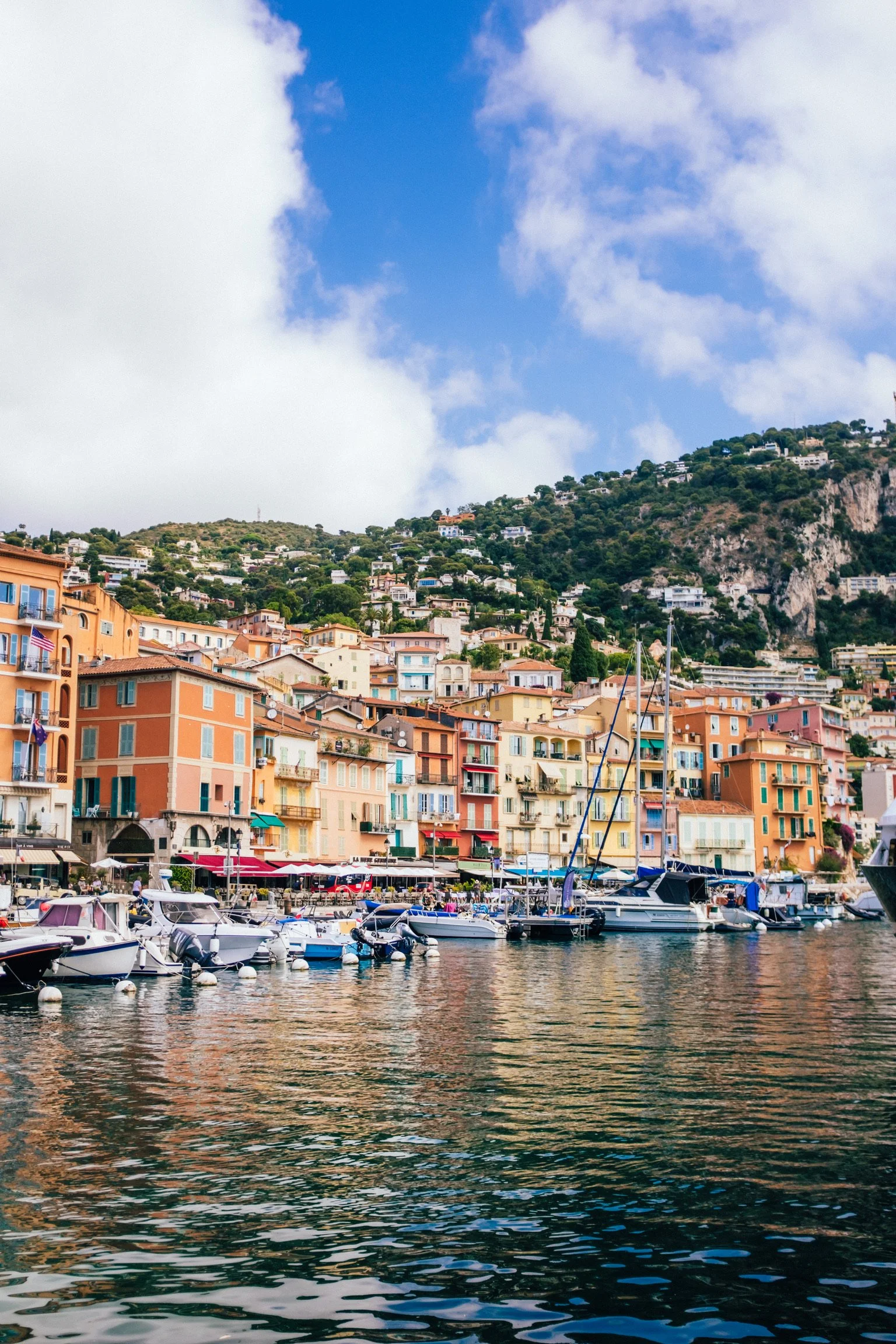 Colorful buildings along a waterfront with boats docked, hillside in the background, and partly cloudy sky.