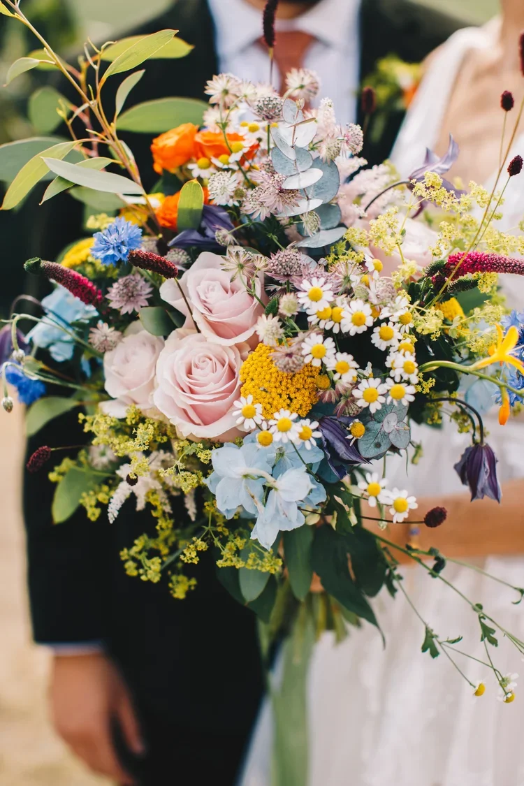 Colorful bouquet of mixed flowers including roses, daisies, and various wildflowers, being held by two people at a celebration or event.