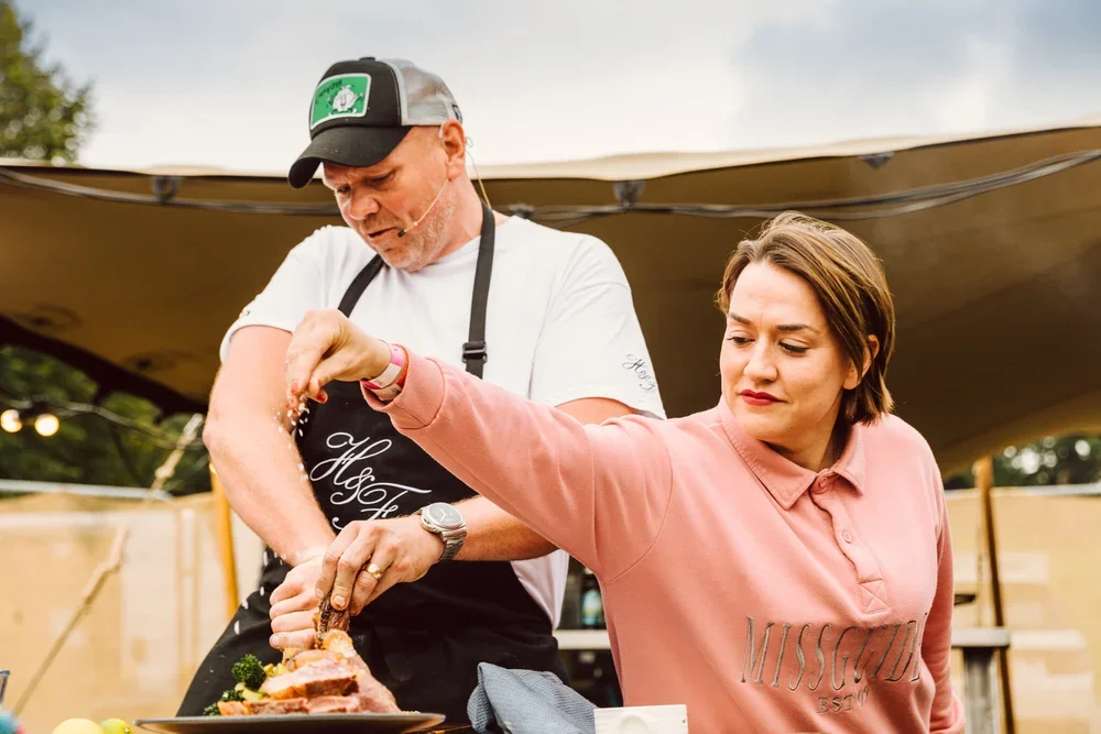 A woman in a pink shirt and a man in a black apron are preparing food outdoors, with the woman sprinkling salt onto a platter of roasted meat.