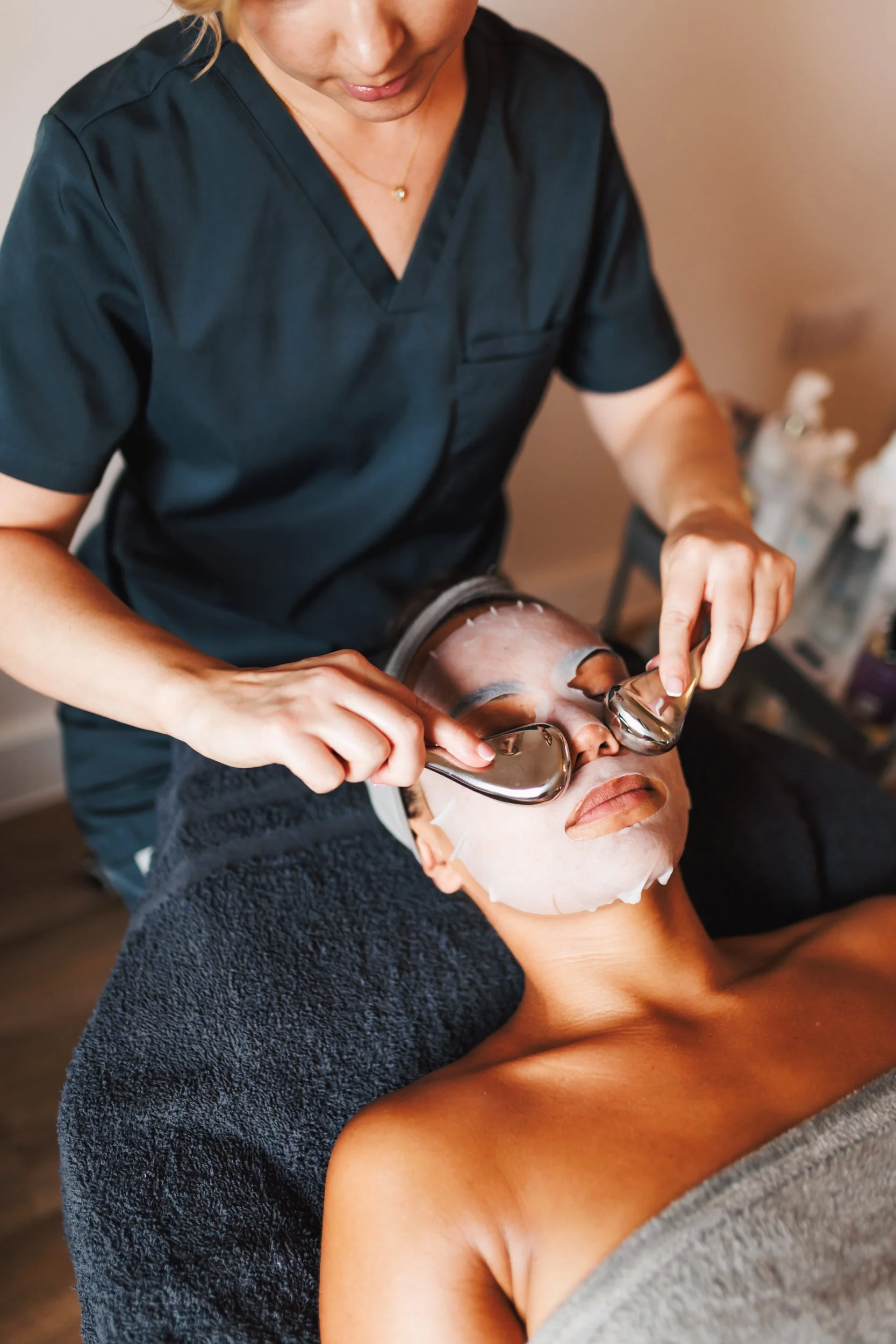 A woman is receiving a facial treatment, with a skincare professional placing cooling gel eye masks on her face, while lying on a treatment bed covered with a towel.
