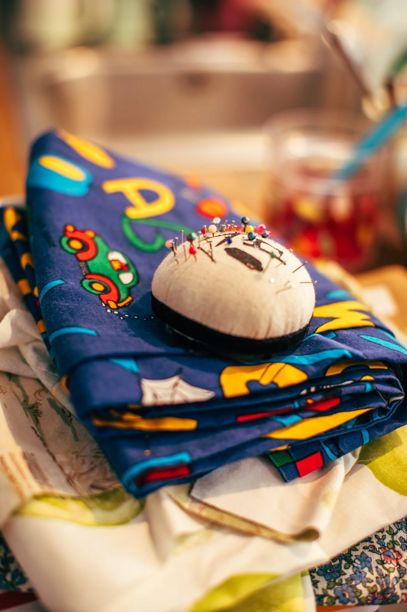 A folded colorful fabric with a cartoon character on it and a small sewing pin cushion on top of it, surrounded by other fabrics and sewing supplies.