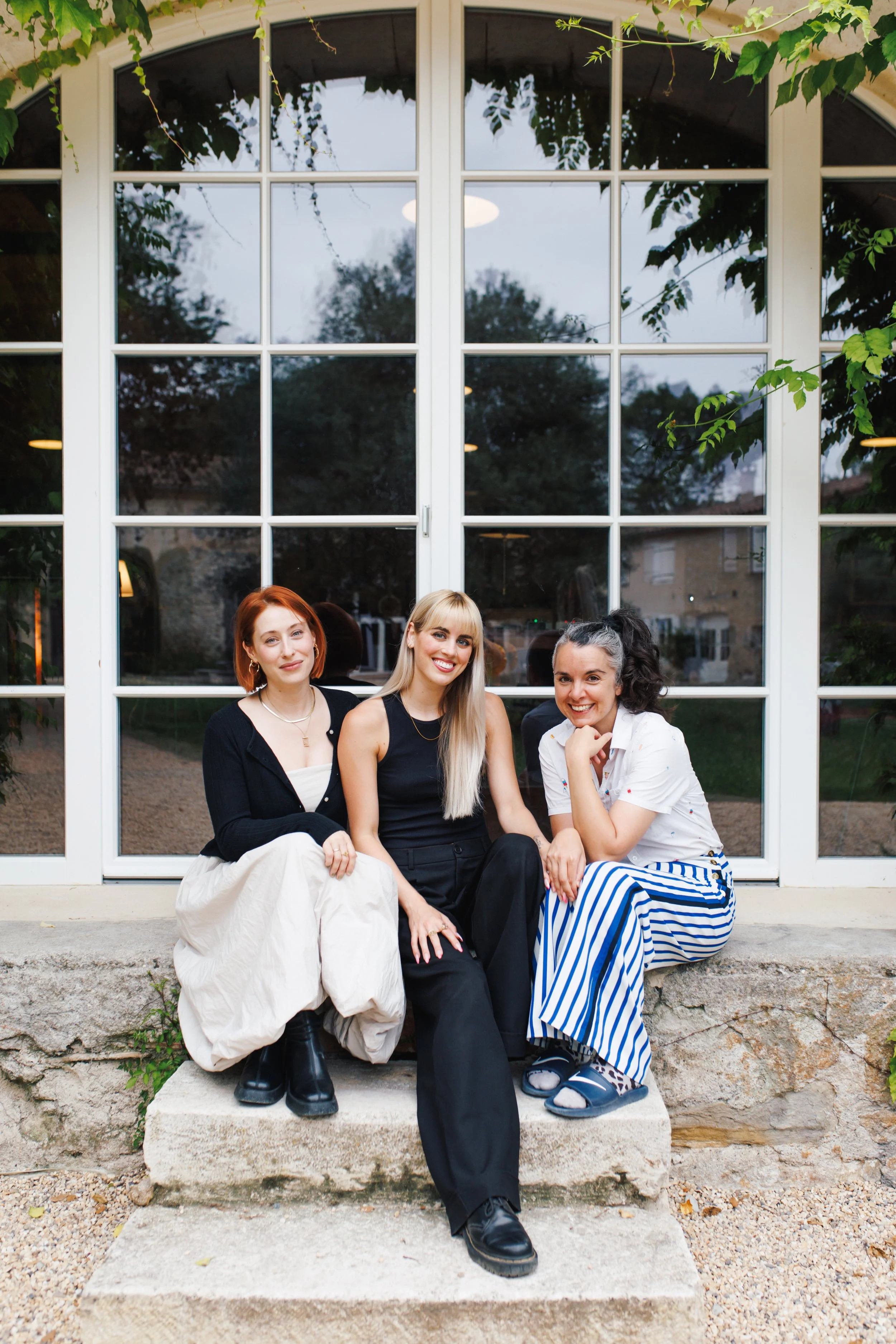 Three young women sitting on a stone step outside near a large window, smiling, with trees and a building reflected in the window.
