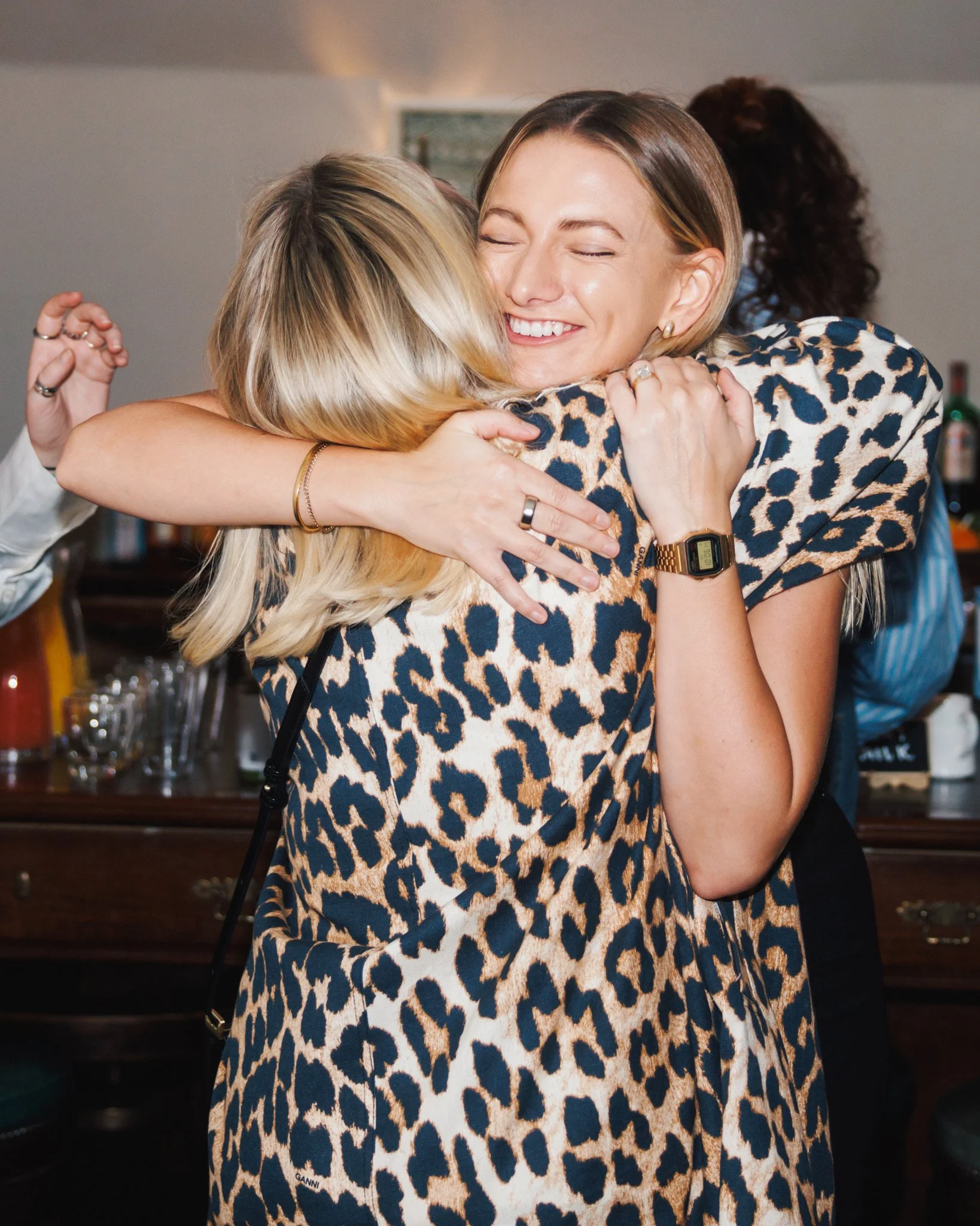 Two women hugging and smiling at a social gathering, with a woman wearing a leopard print dress and a gold watch.