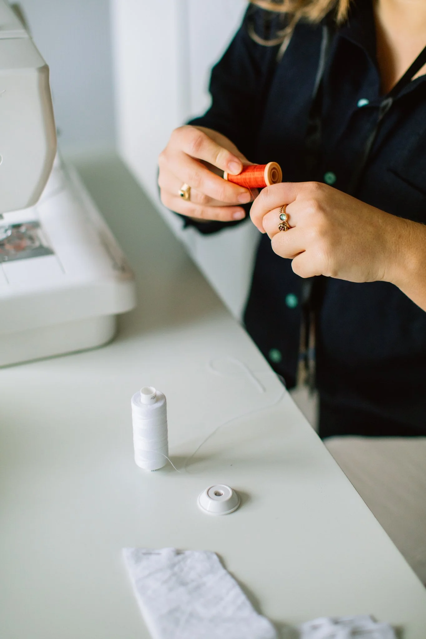 Person threading a needle with white thread, sewing to a small fabric piece on a white table, sewing supplies including a spool of white thread and a thimble.