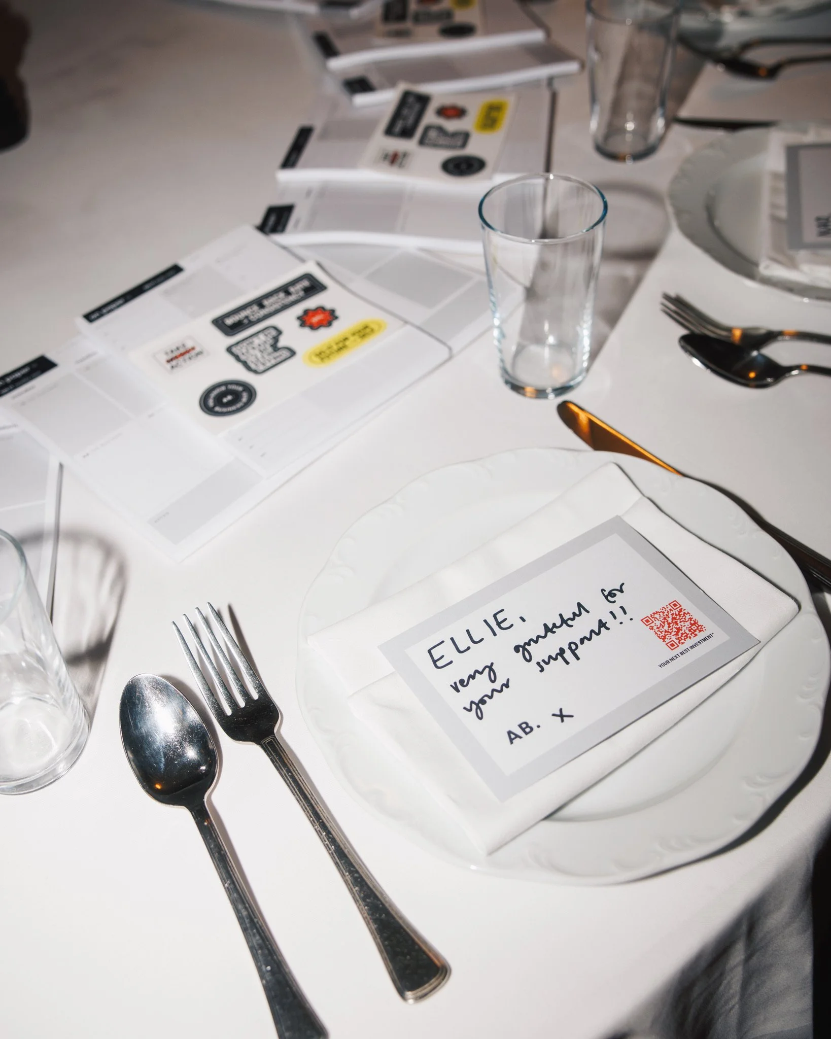 A banquet table setting with white dinnerware, a folded napkin with a handwritten note, and printed program brochures. The note reads, 'ELLIE, very happy for your moment to shine!! AB. x' and has a red QR code. Silverware and empty glasses are arrang