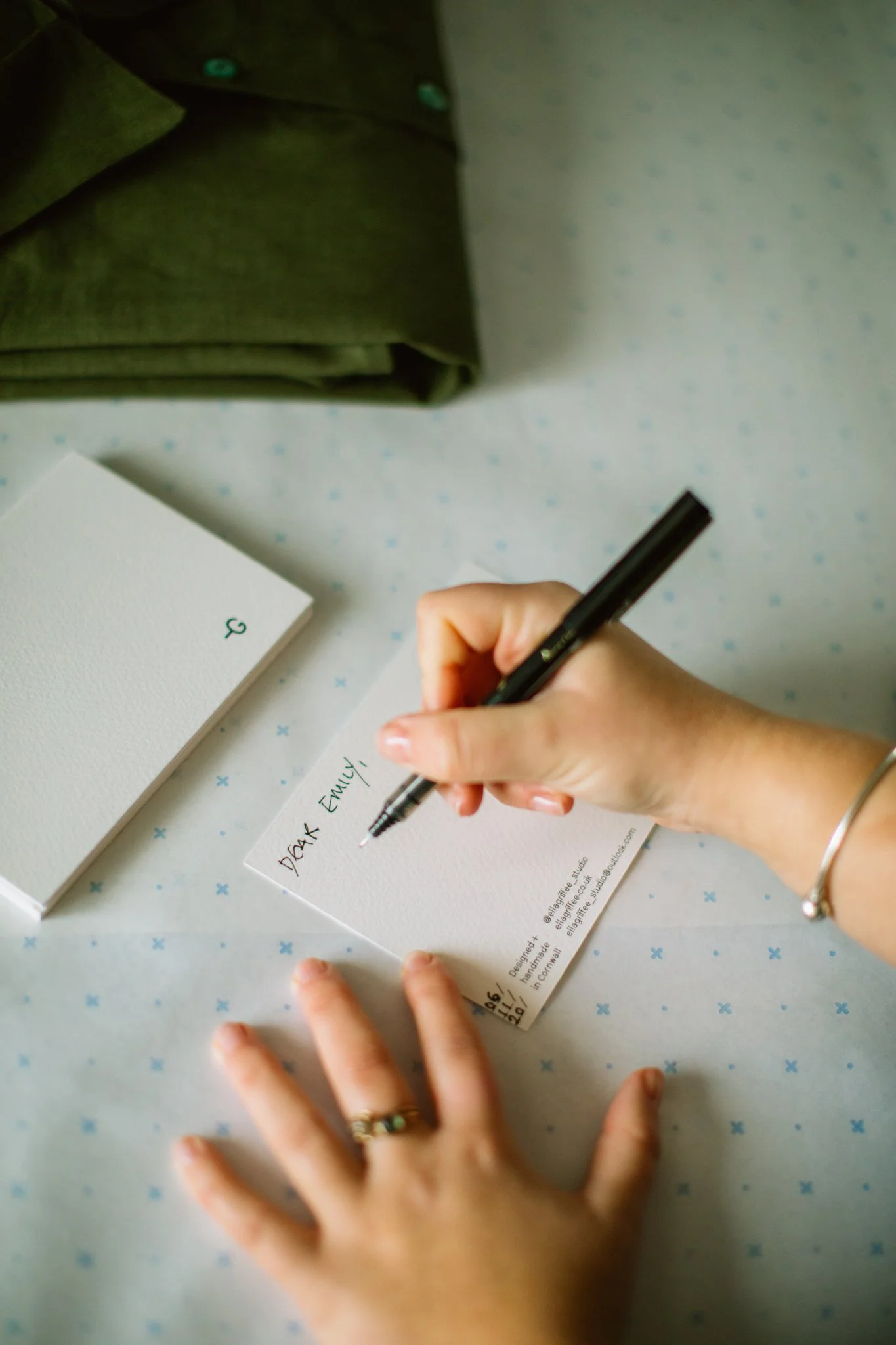 A person writing a note that says 'Dear Emily' on a small white card, with a green bag and a white notebook nearby on a table with a light blue patterned tablecloth.