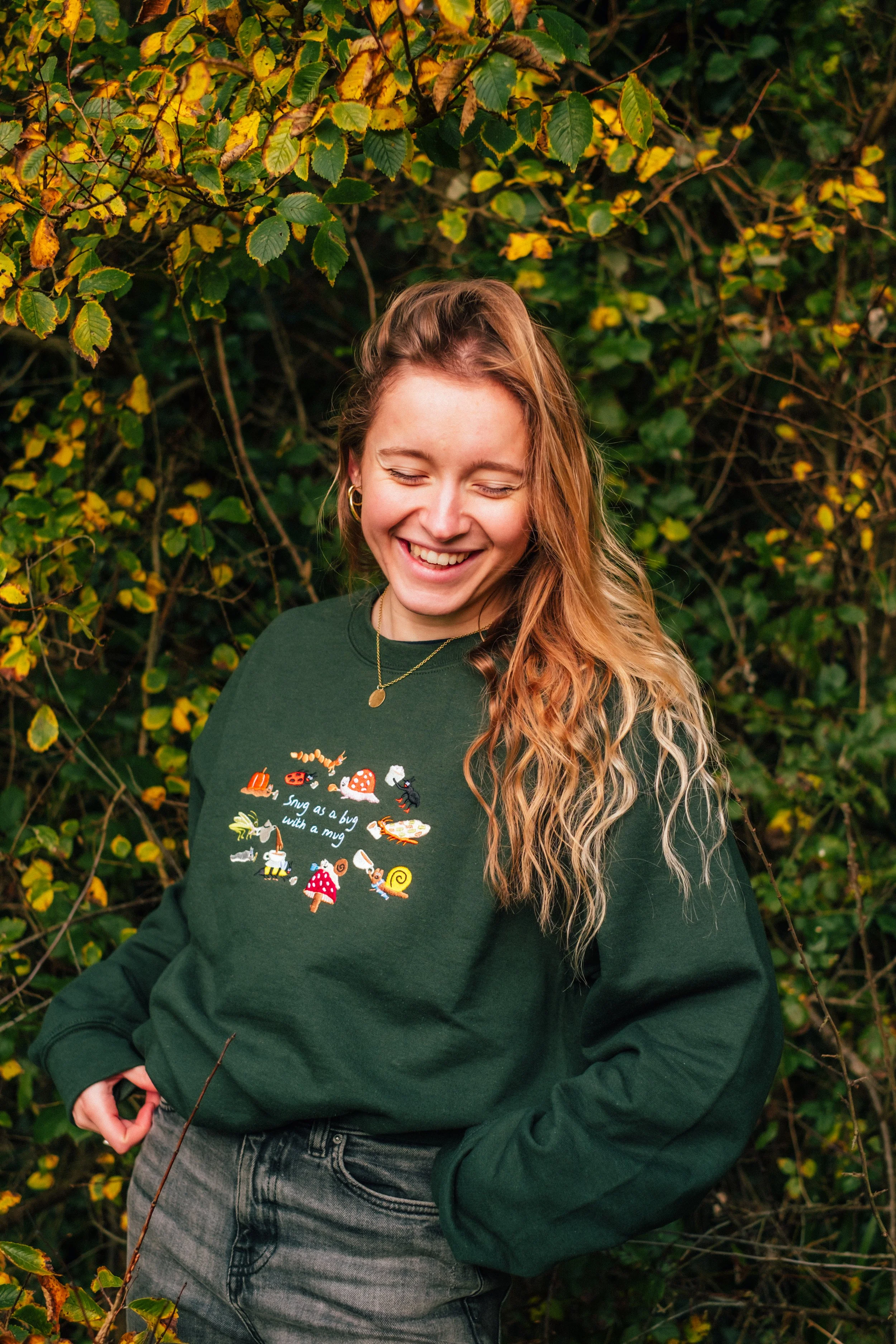 A young woman smiling with her eyes closed, wearing a green sweatshirt with colorful bug and mushroom illustrations, standing outdoors in front of leafy yellow and green foliage.