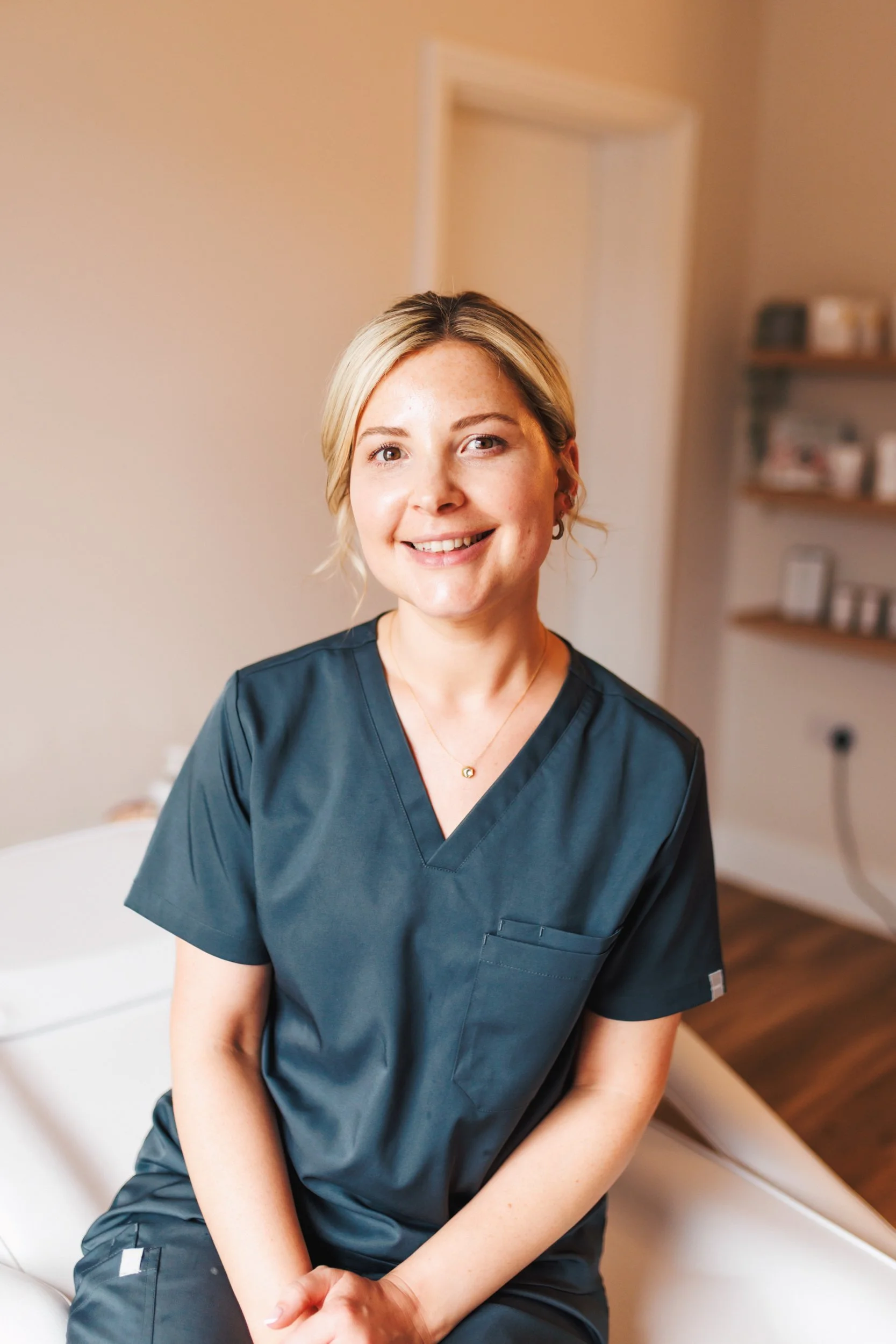 A smiling woman in medical scrubs sitting on a medical bed in a clinical setting.