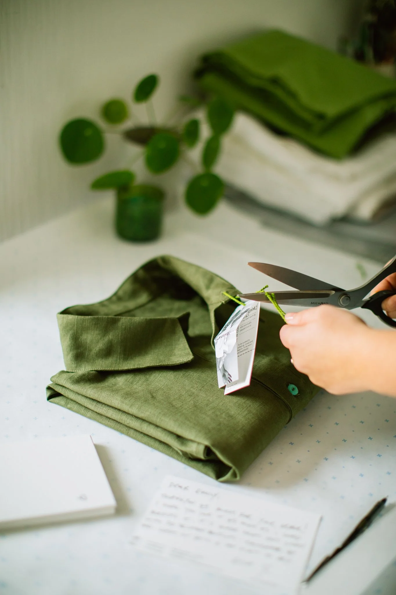 Person cutting a tag off a folded green shirt with scissors on a white table, with a notepad, a pen, folded fabric in the background, and a small potted plant.