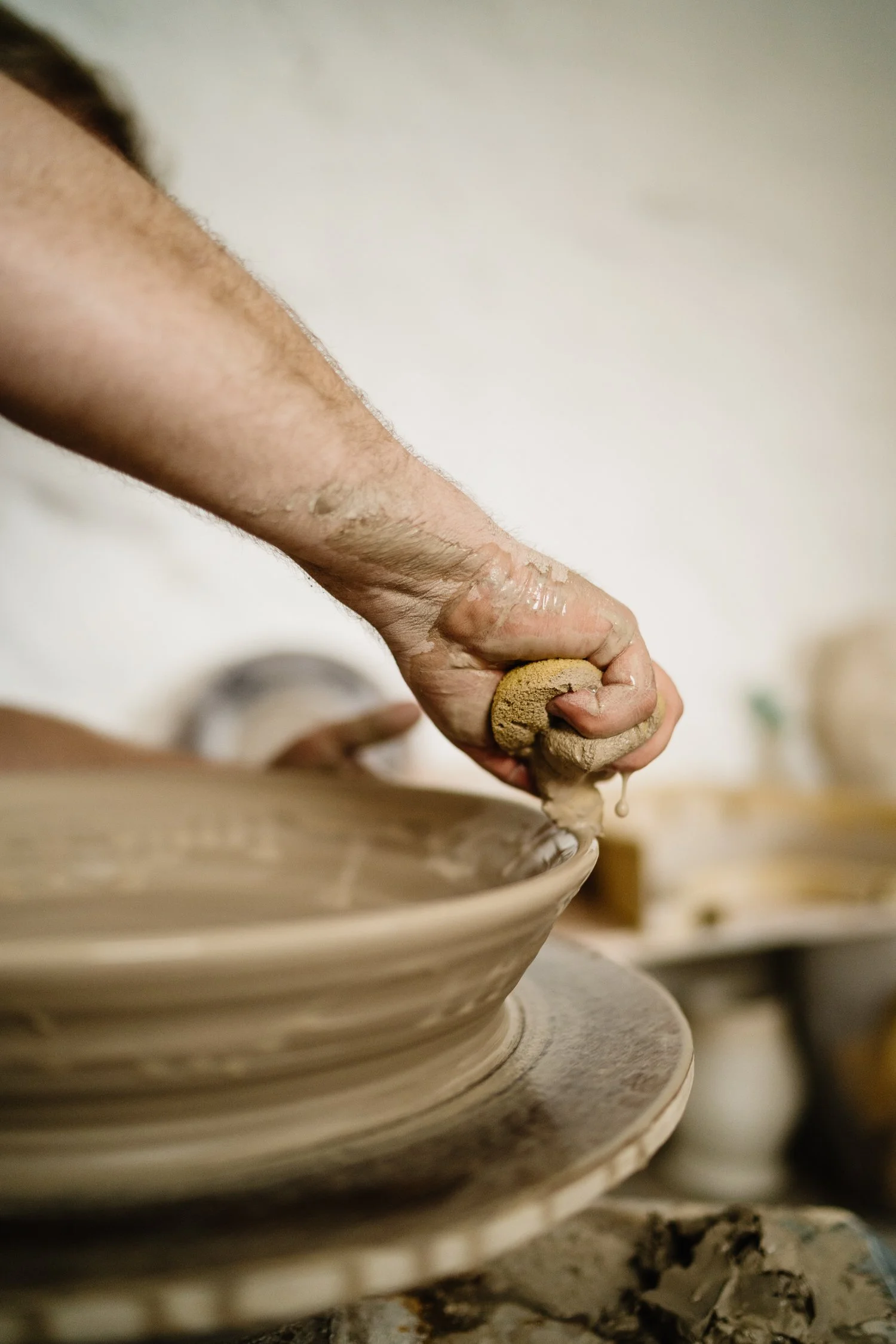 A person shaping clay on a pottery wheel with their hand, focusing on the earthy clay and their hand covered in slip.