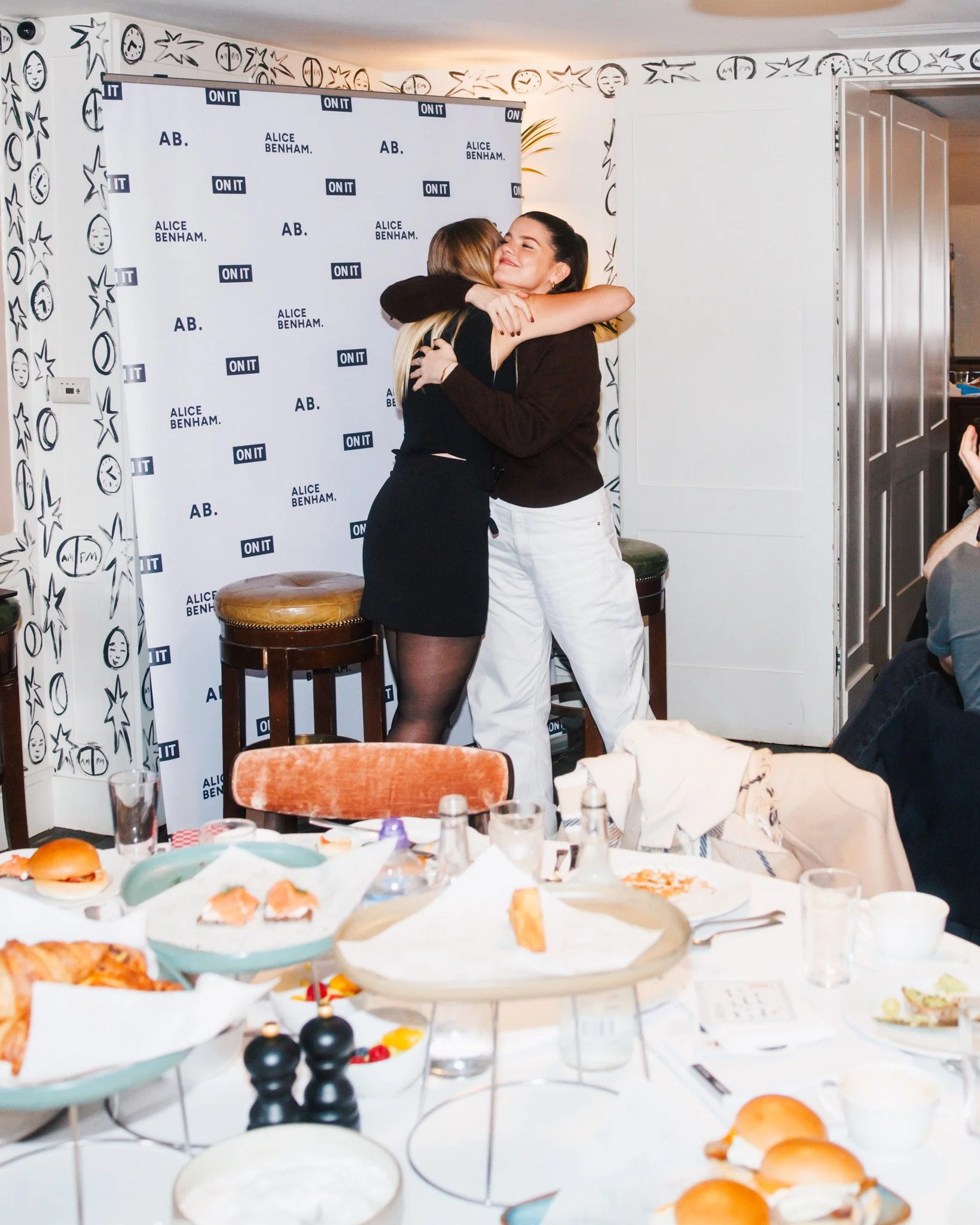 Two women hugging on stage at an event, with a branded backdrop reading 'ALICE BENHAM' and 'ON IT' behind them. A table in front is set with food, drinks, and tableware for a meal, indicating a celebratory or social gathering.