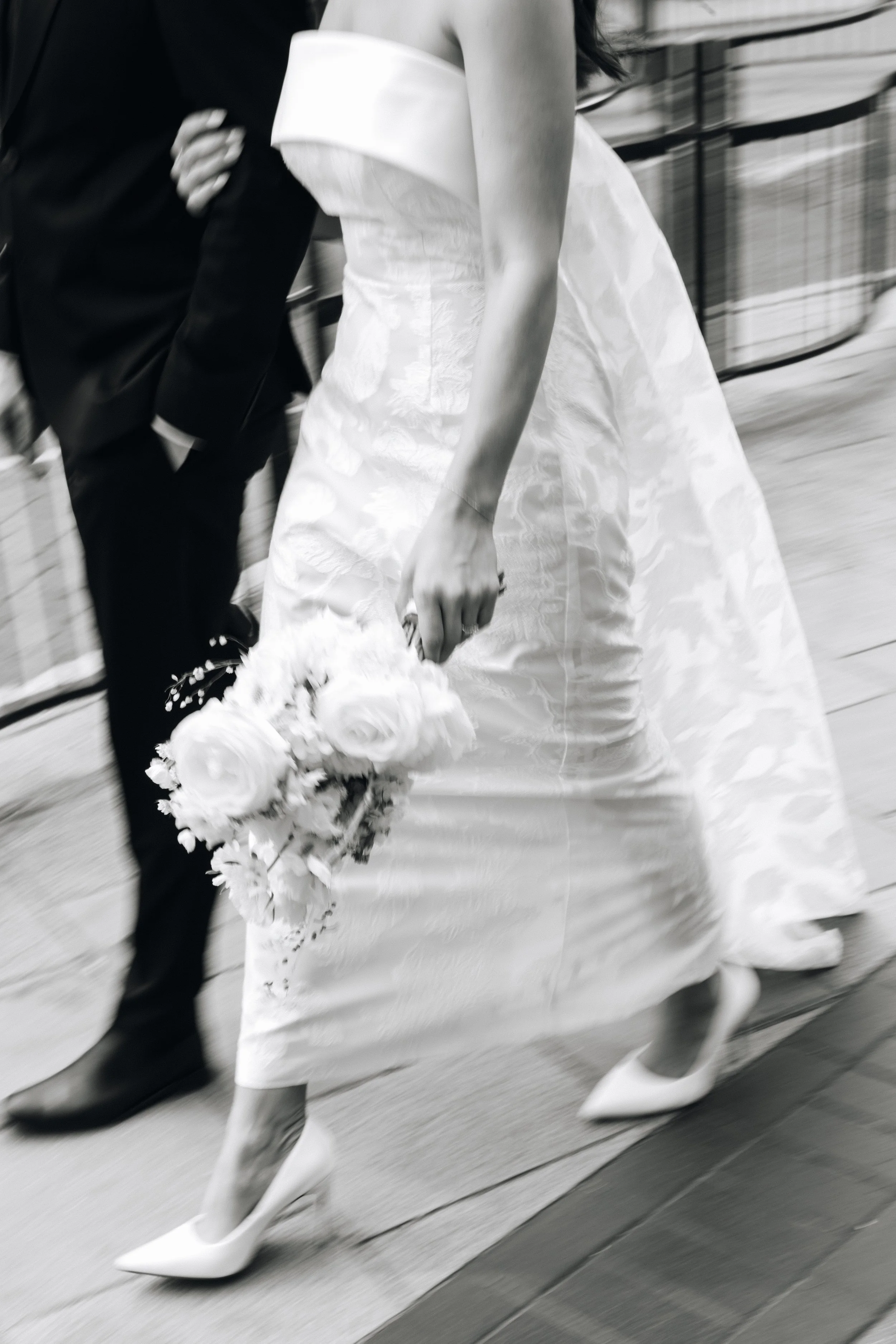 A woman in a wedding dress holding a bouquet of flowers, walking with a man in a suit, both captured in motion.