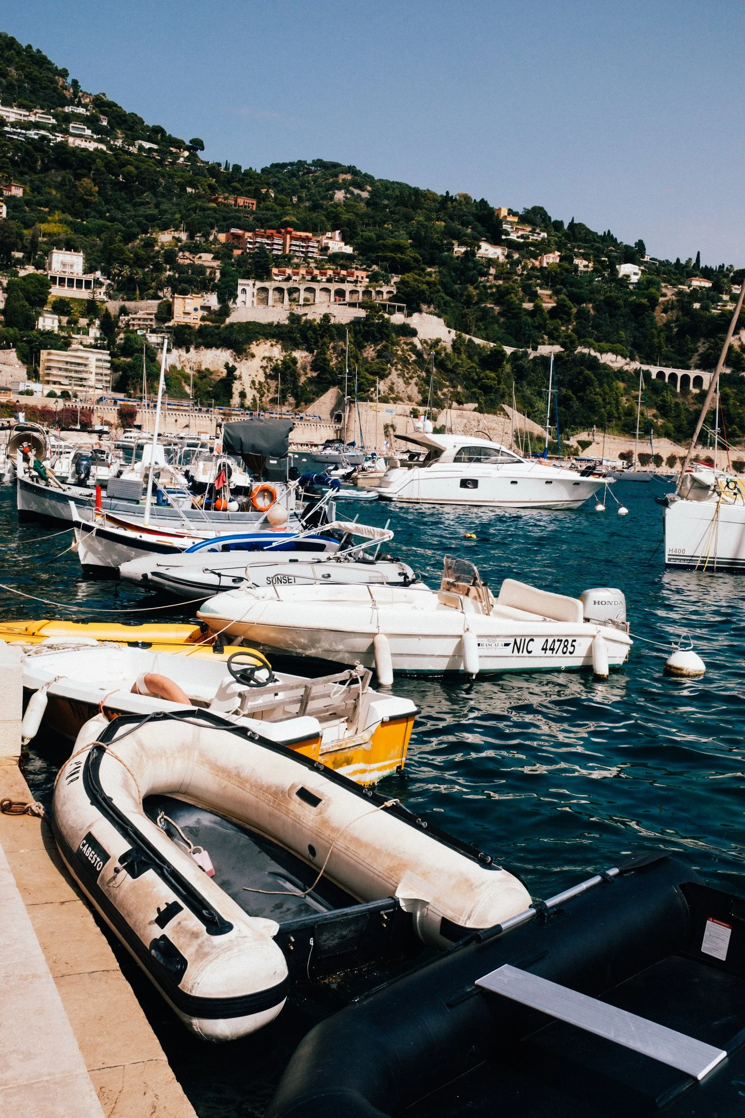 Marina with boats docked, hillside with houses in the background, clear blue sky.