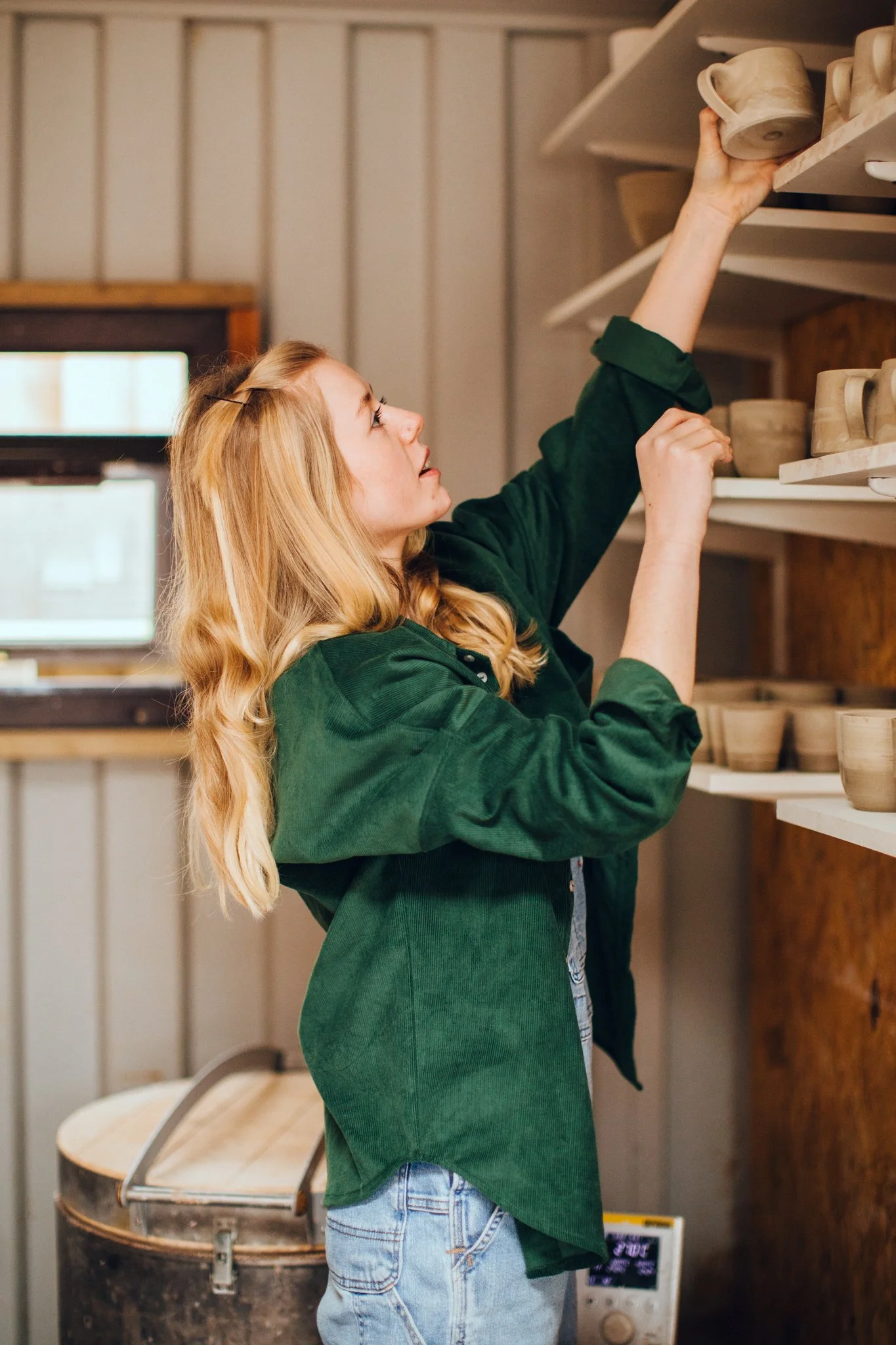 A woman with long blonde hair, wearing a green jacket and jeans, reaches up on a wooden shelf to pick up a ceramic mug in a pottery shop.