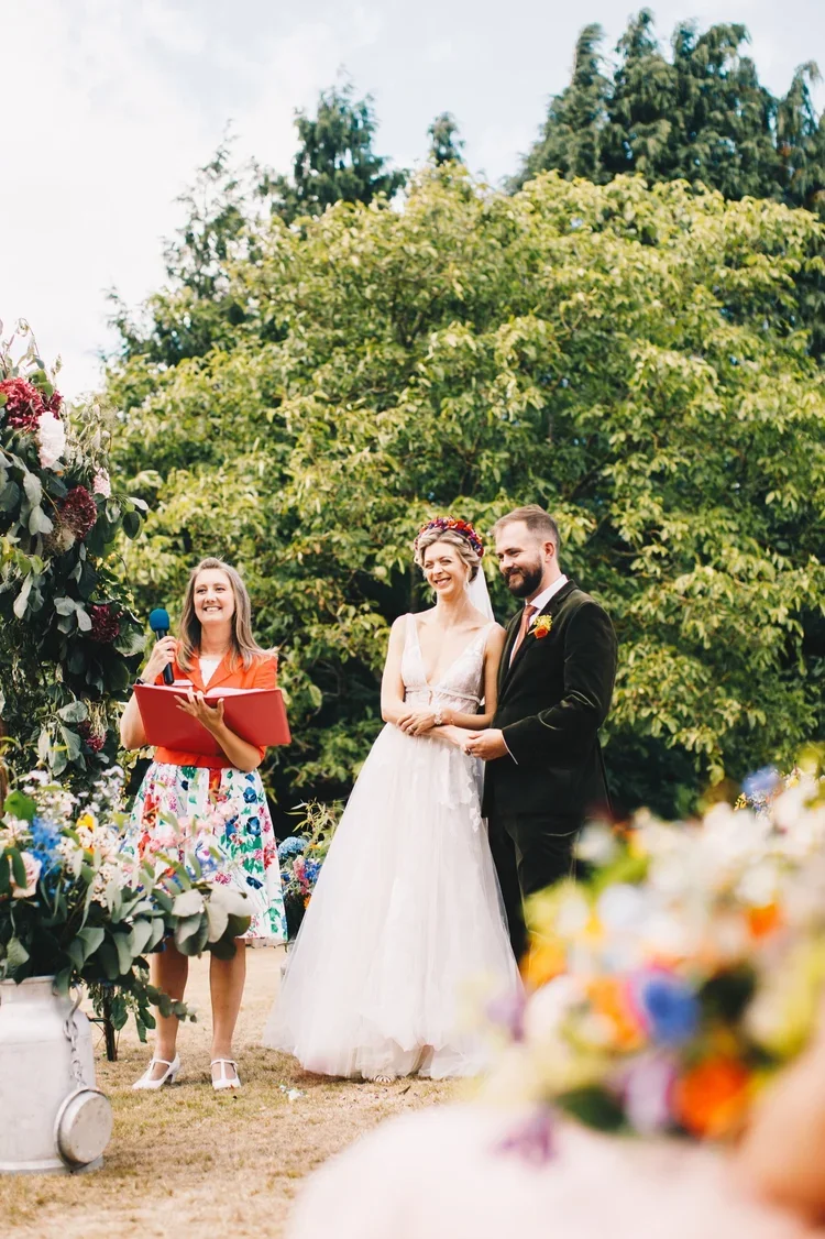 A wedding ceremony taking place outdoors with a bride and groom standing together, smiling, while a woman reads from a book, holding a microphone. The scene is lush with green trees and colorful flowers.