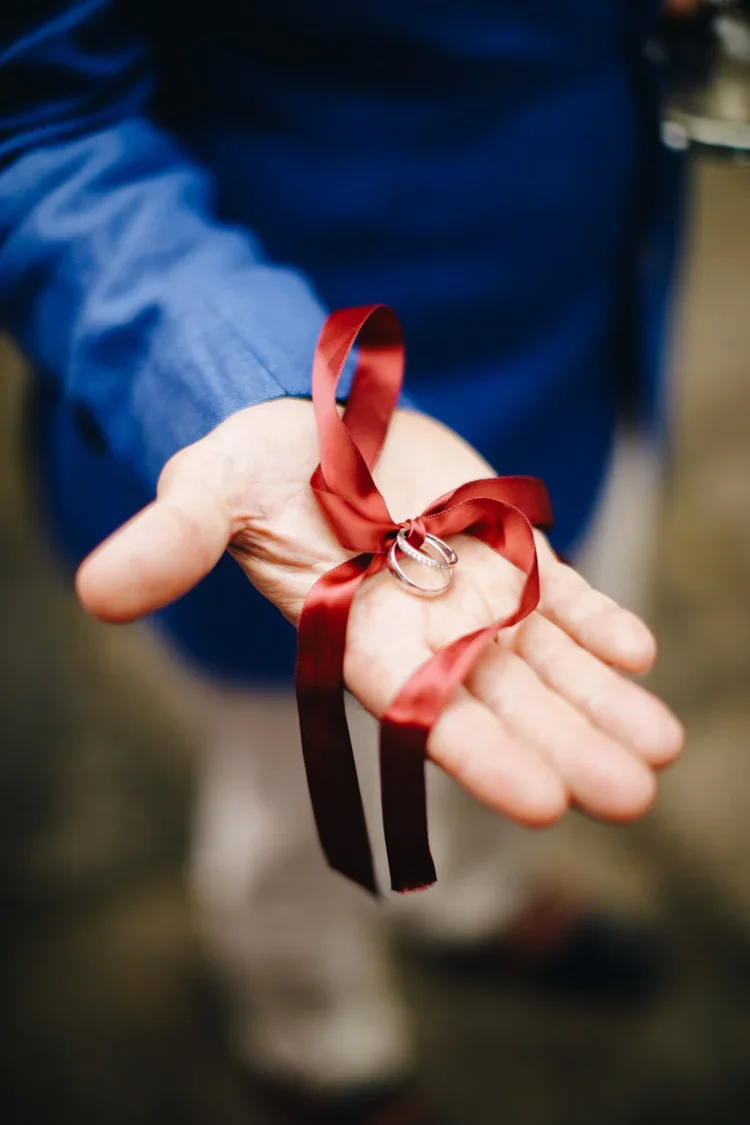 A person in a blue shirt holds a red ribbon with wedding rings attached, symbolizing marriage.