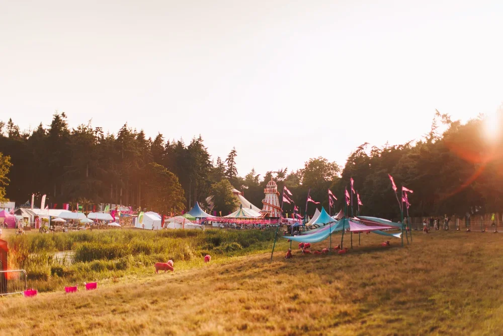A fairground with colorful tents, flags, and a large carousel with a striped top, set against a backdrop of trees at sunset.