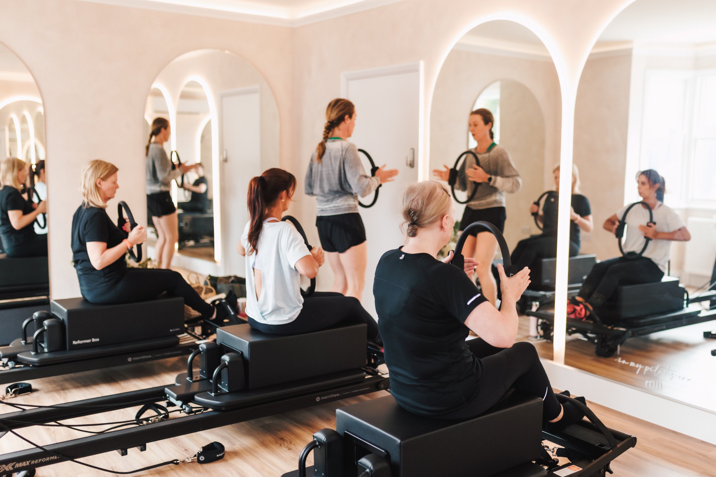 A group of women participating in a Pilates class using reformer machines in a bright, mirrored studio.