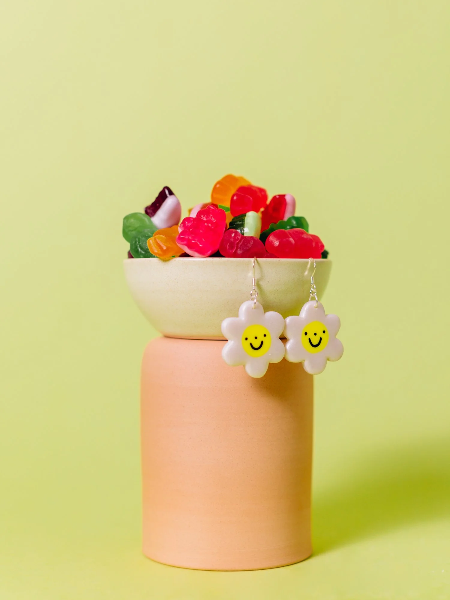 Colorful gummy bears in a yellow bowl, placed on a pink stand, with white flower-shaped earrings featuring happy face icons hanging from the bowl.