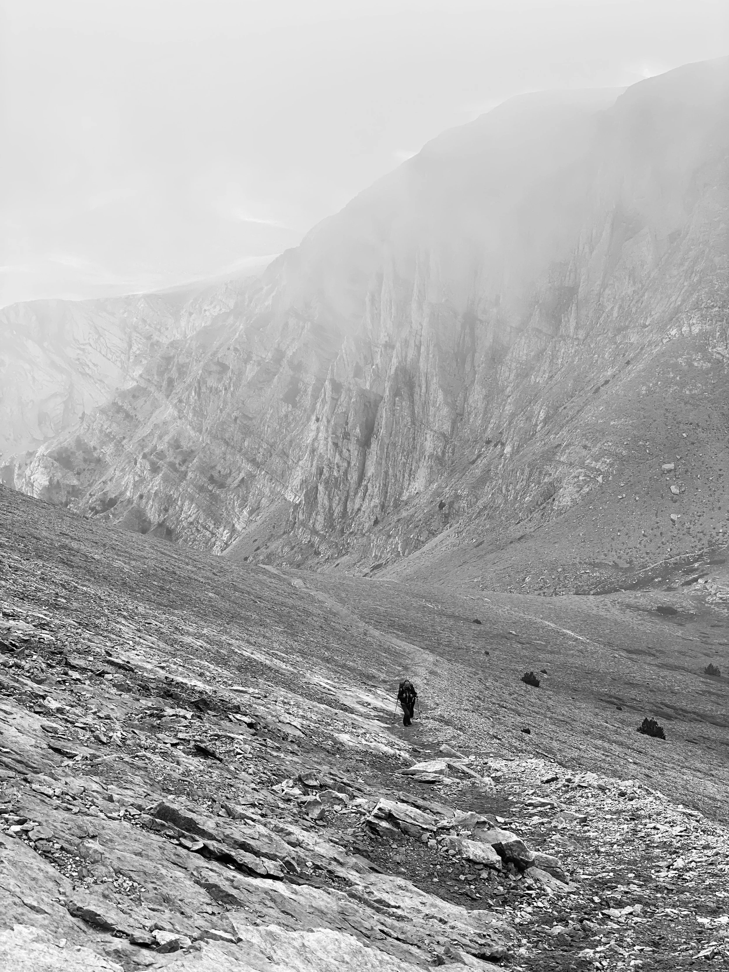 A black and white photo of a hiker walking on a rocky trail up Mt. Olympus in Greece in a vast, mountainous landscape with steep cliffs and foggy sky.