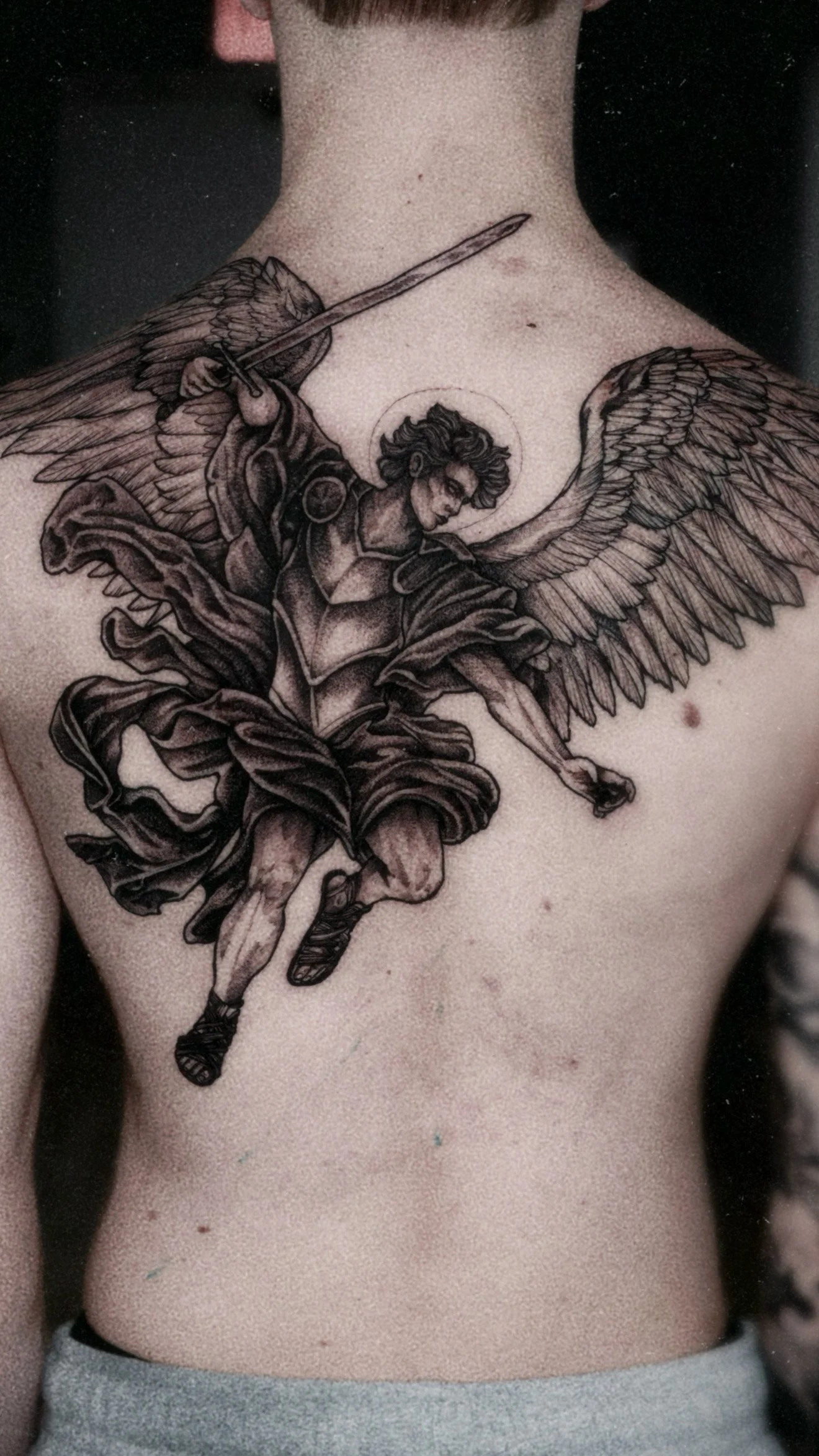 A large, neoclassical style back tattoo of the Archangel Michael in armor with expansive wings, wielding a sword in a dynamic, mid-air pose.