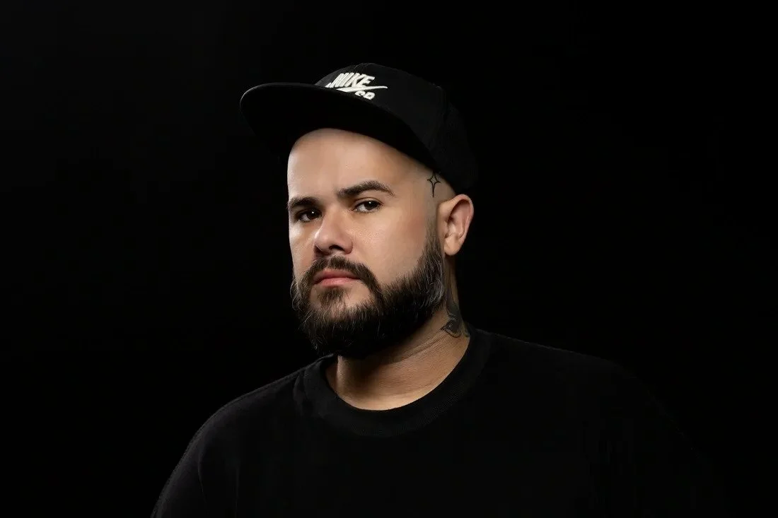 Portrait of a man with a beard and tattoos wearing a black Nike cap and black shirt against a black background.