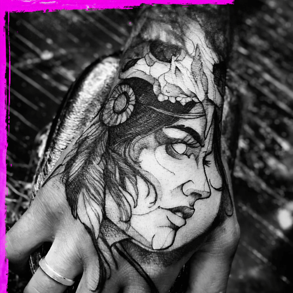 Black and white photograph of a tattoo on a person's hand, featuring a woman's face with long hair, a seashell, and a skull, with a wooden background.