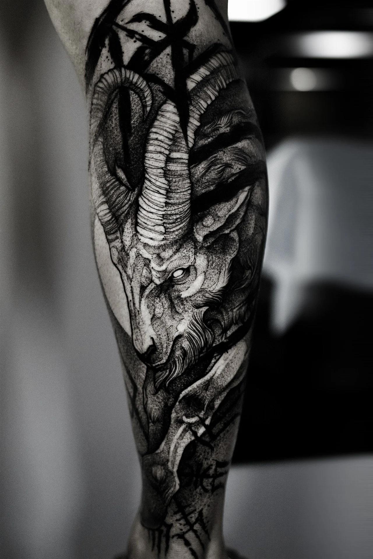 A striking black-and-grey tattoo on a person's calf featuring a majestic ram or goat head. The design uses heavy contrast, intricate stippling on the skin, and bold, illustrative linework on the large curved horns.