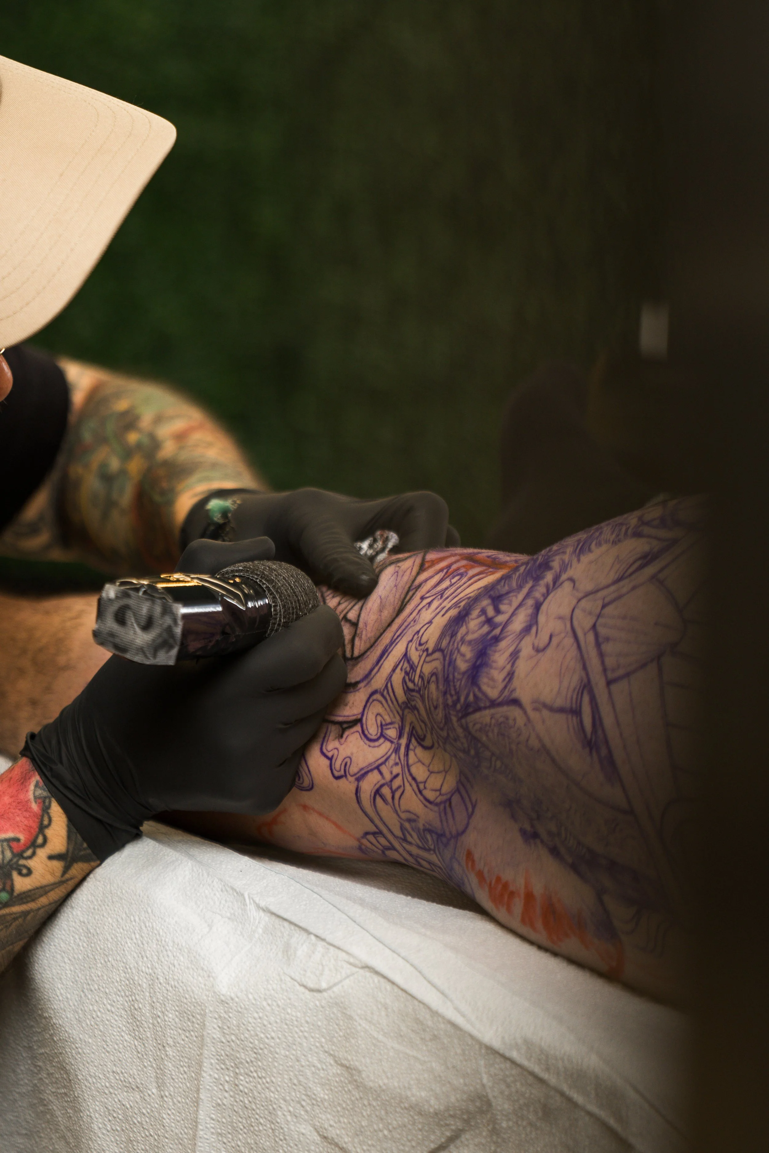 A person getting a tattoo on their arm by a tattoo artist. The artist is using a tattoo machine and wearing black gloves. The person's arm has a partially completed tattoo with visible outline design.