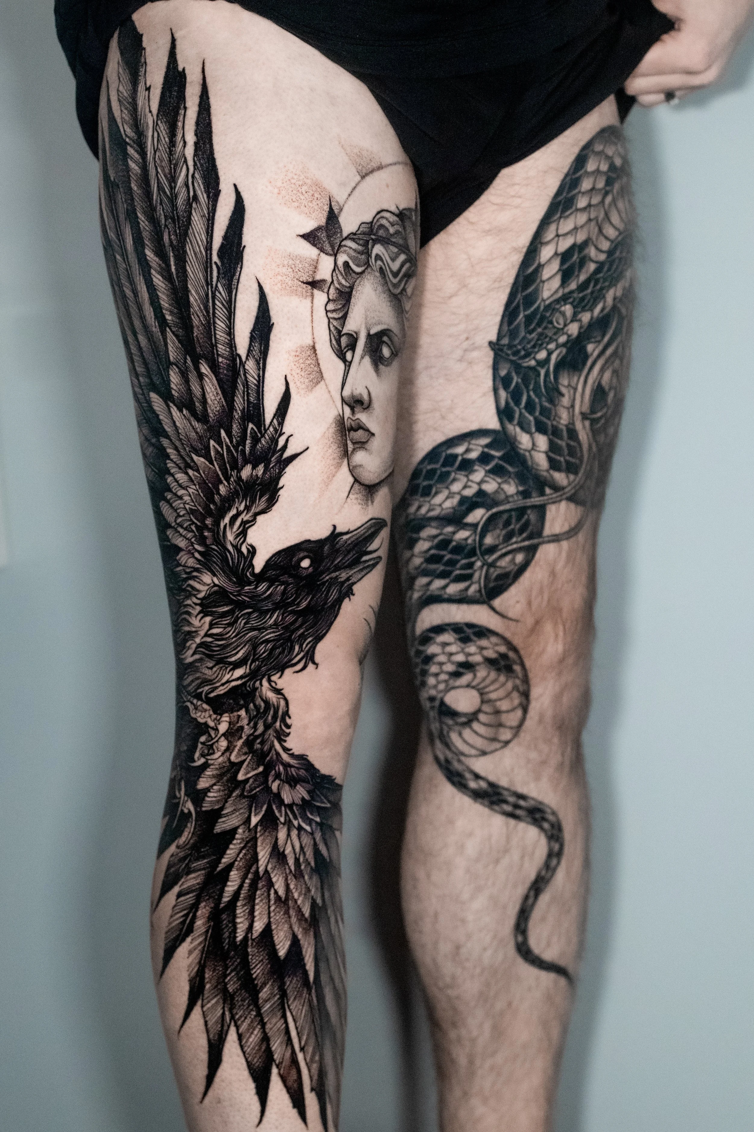 Black-and-gray raven and serpent leg tattoos.