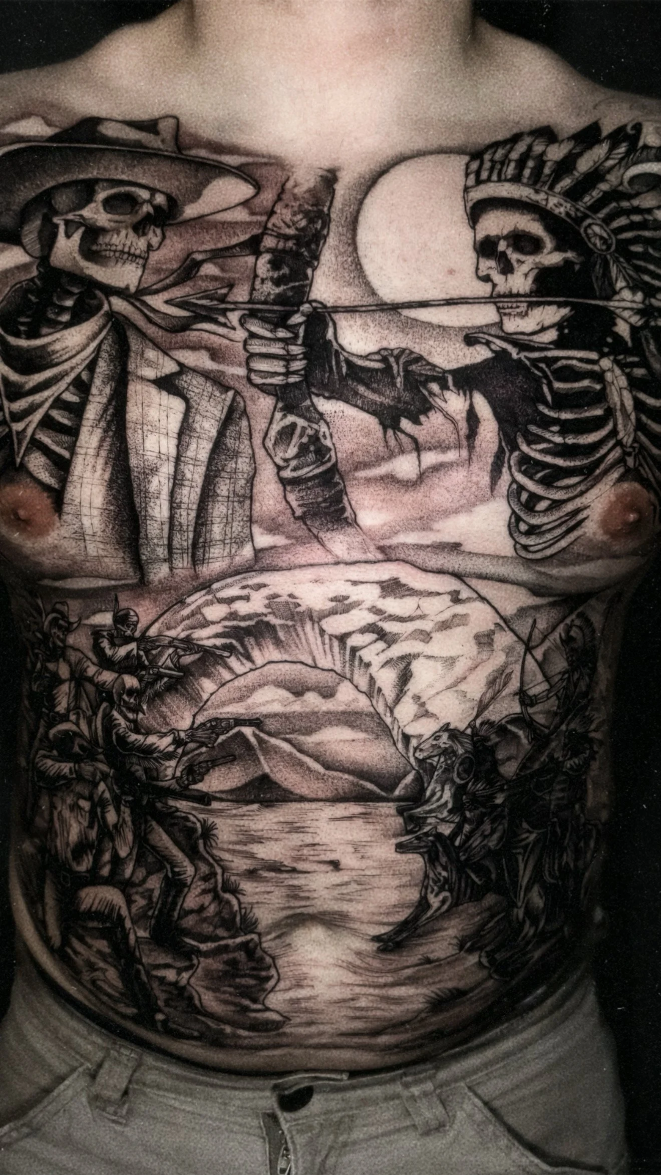 An expansive torso tattoo depicting a "Wild West" scene with skeletal figures: a cowboy skeleton and a Native American skeleton face off at the top, while a battle scene unfolds across the stomach.
