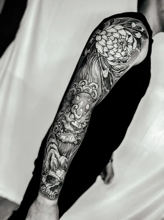 Detailed black and gray tattoo sleeve featuring a fierce lion's face, a skull, and a large flower on a person's arm.