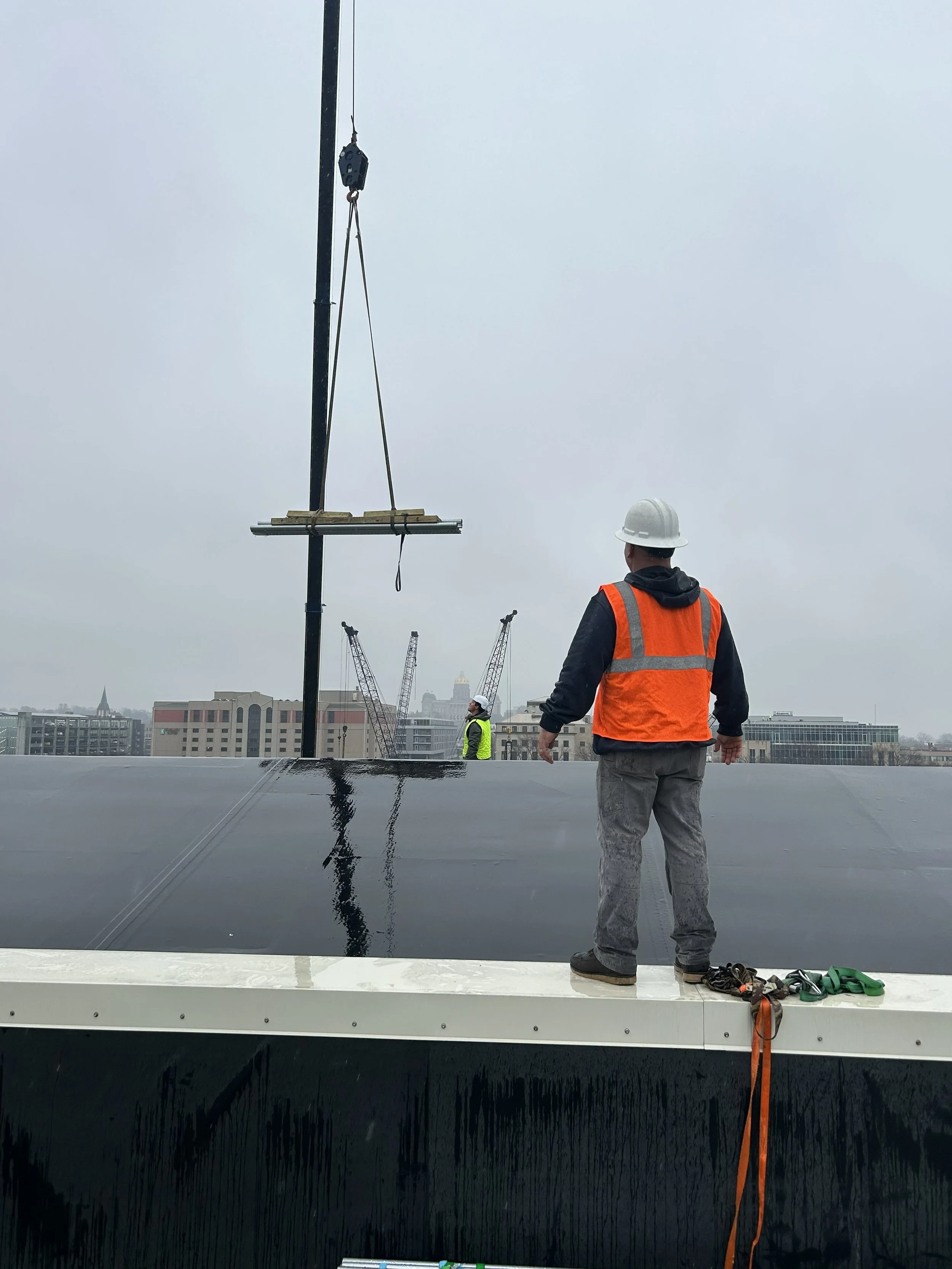 Construction worker wearing a white hard hat and orange safety vest standing on a rooftop, with tools on the edge, overlooking a cityscape with cranes in the background.