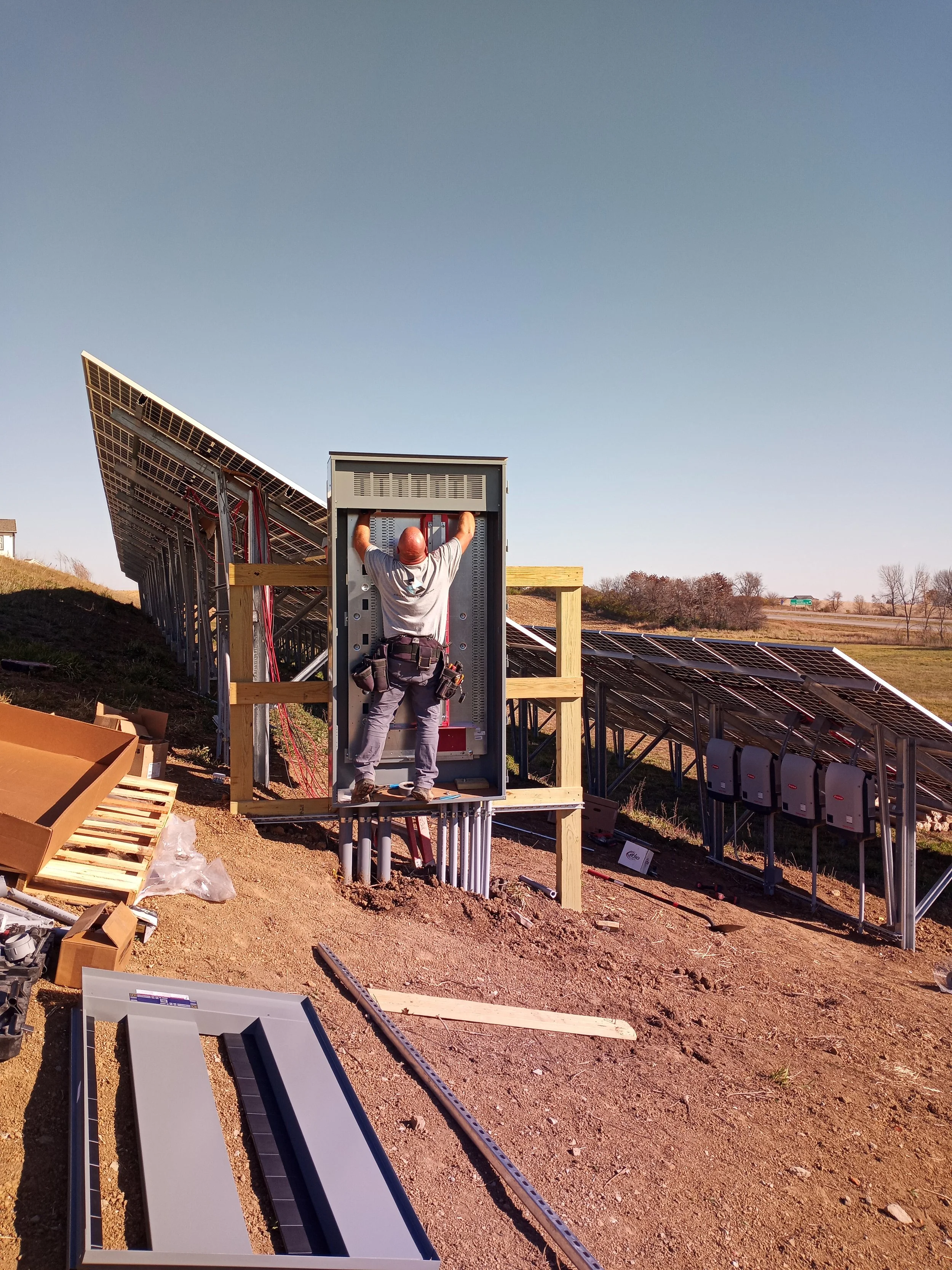 Worker installing or repairing electrical components inside a large outdoor electrical panel or box near solar panels on a sunny day.