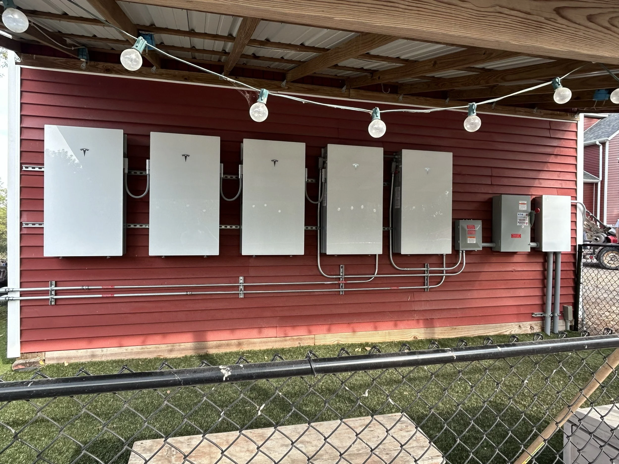 Multiple electrical boxes and panels mounted on a red house exterior wall, with wiring and conduit beneath, and a string of outdoor bulbs hanging above.