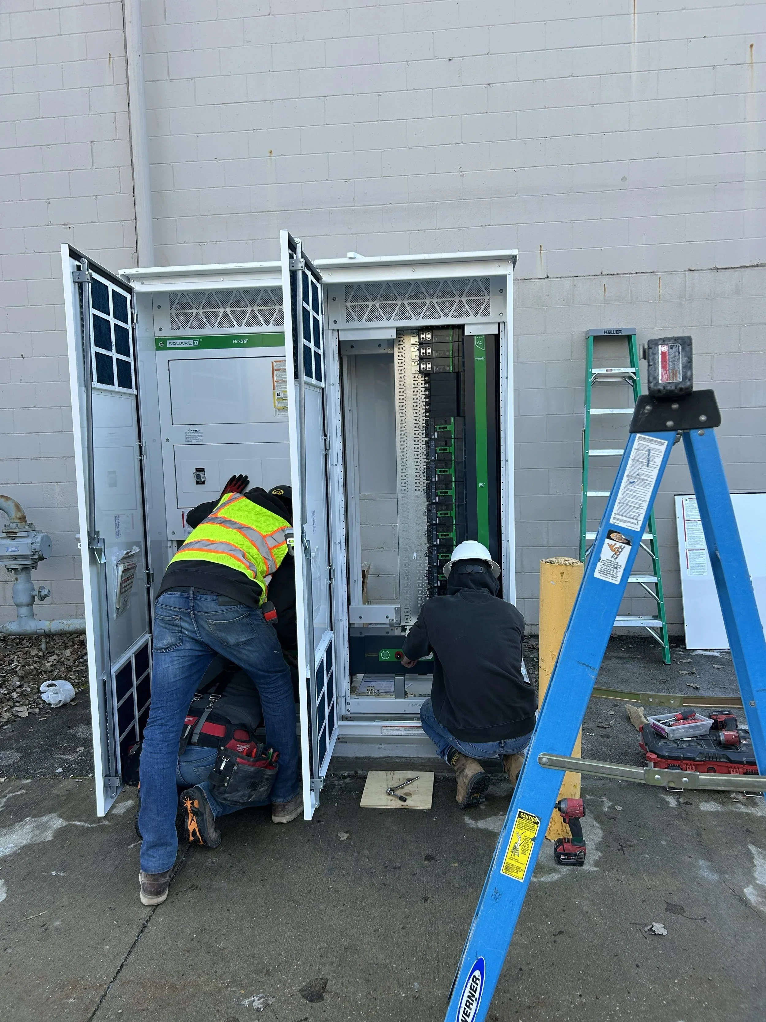 Two workers installing or repairing an electrical panel outside a building, with ladders and tools nearby.