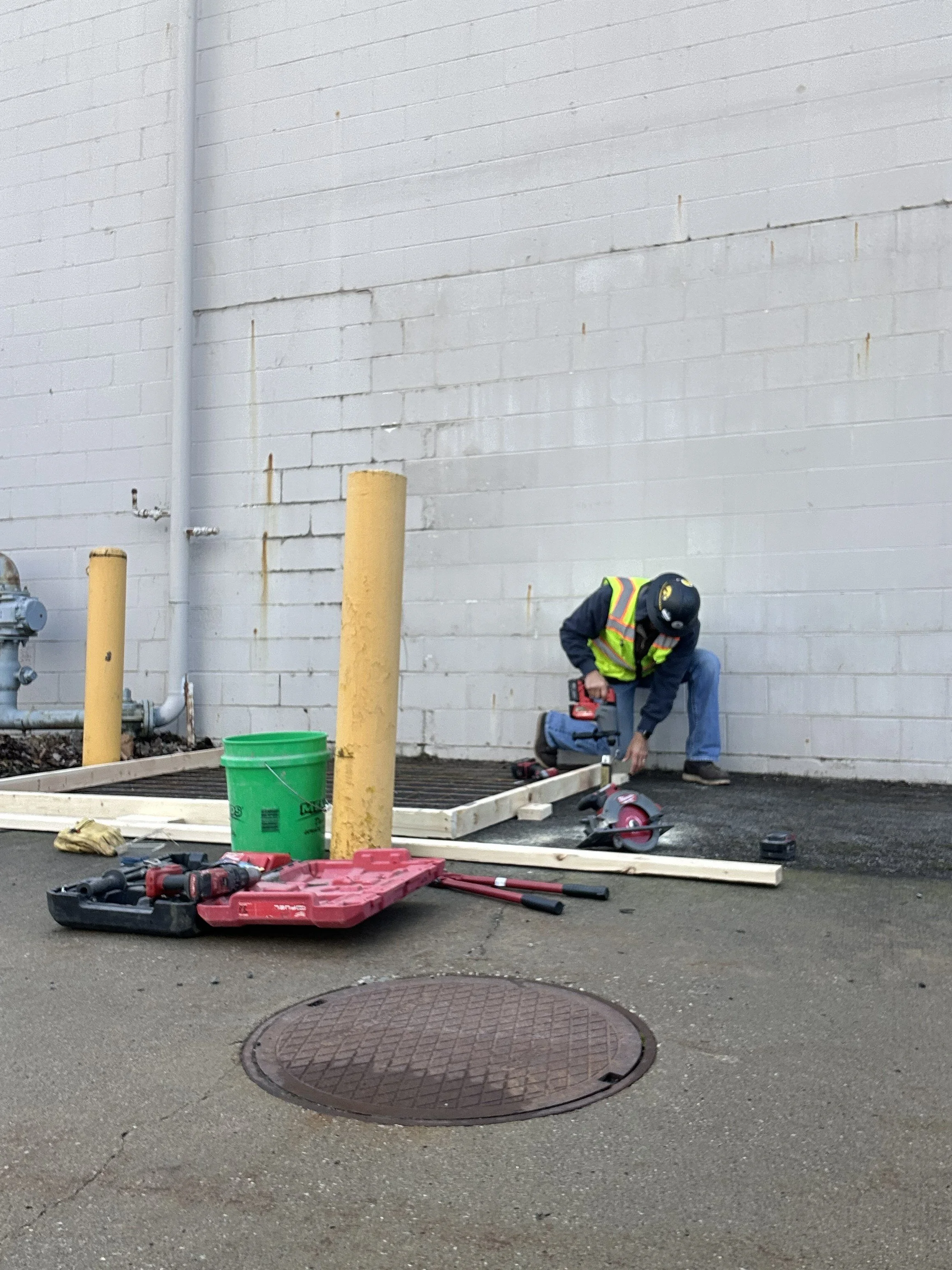 A construction worker wearing a safety vest and helmet is working on a project outdoors near a white brick wall. The worker is using a power tool on a wooden frame or small structure, with various tools and equipment scattered around, including a green bucket, a toolbox, and power saws.