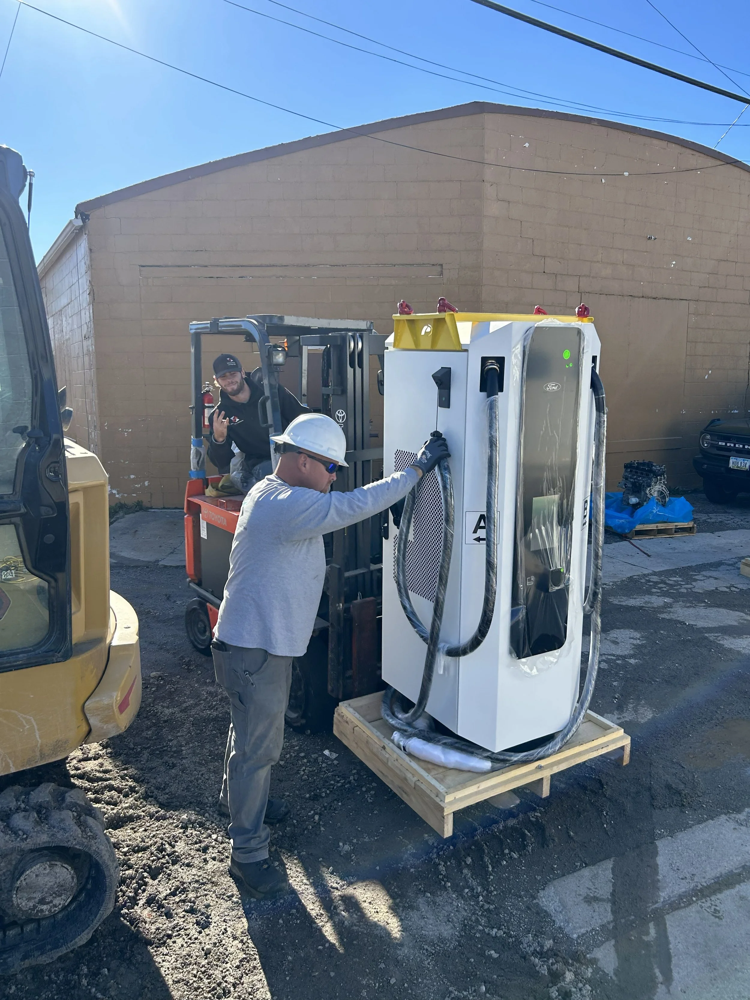 Two men working outdoors with a forklift and a large piece of equipment, one man in a hard hat and gloves inspecting the equipment with a power cord, another man in a cap smiling in the background.